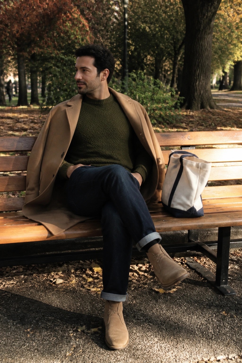Man in tan overcoat and green sweater sits on autumn park bench with tote bag