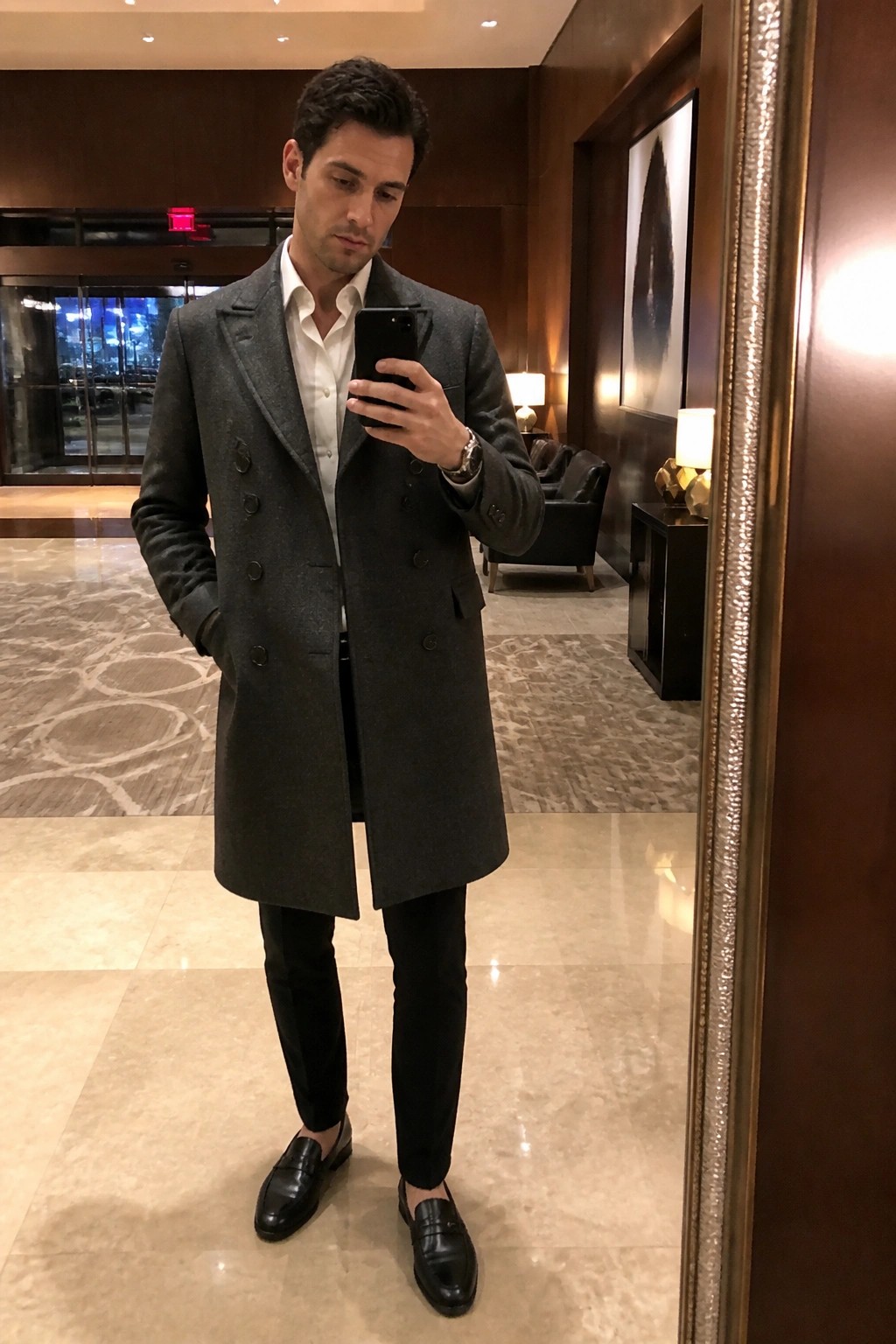 Man wearing a dark gray double breasted wool overcoat over a white collared shirt with black slim trousers and black leather loafers while holding a phone in a mirror selfie.