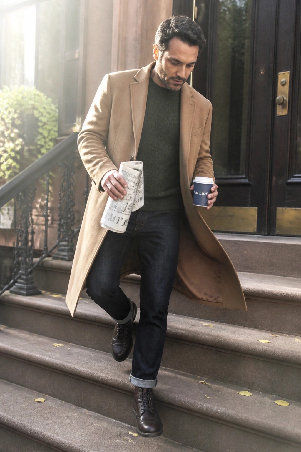 Man in tan coat and green sweater descends stone steps holding coffee and newspaper.