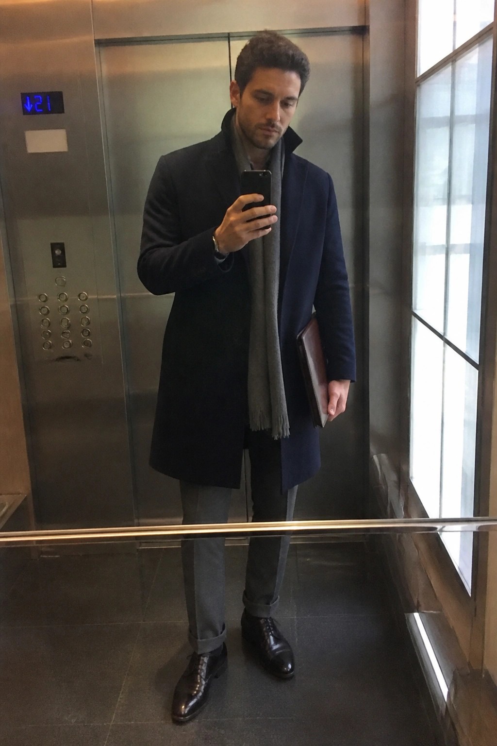 Man in navy overcoat and gray scarf taking mirror selfie in elevator.