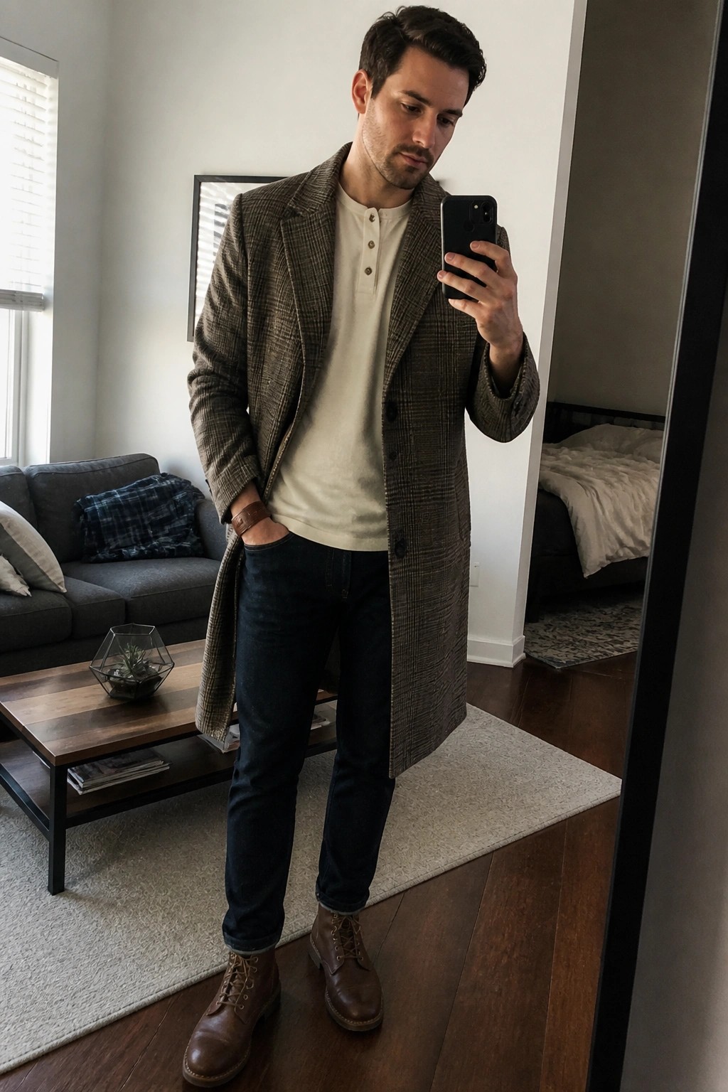 Man wearing a long brown plaid wool overcoat over a cream henley shirt, dark blue jeans, and brown leather boots while taking a mirror selfie indoors.
