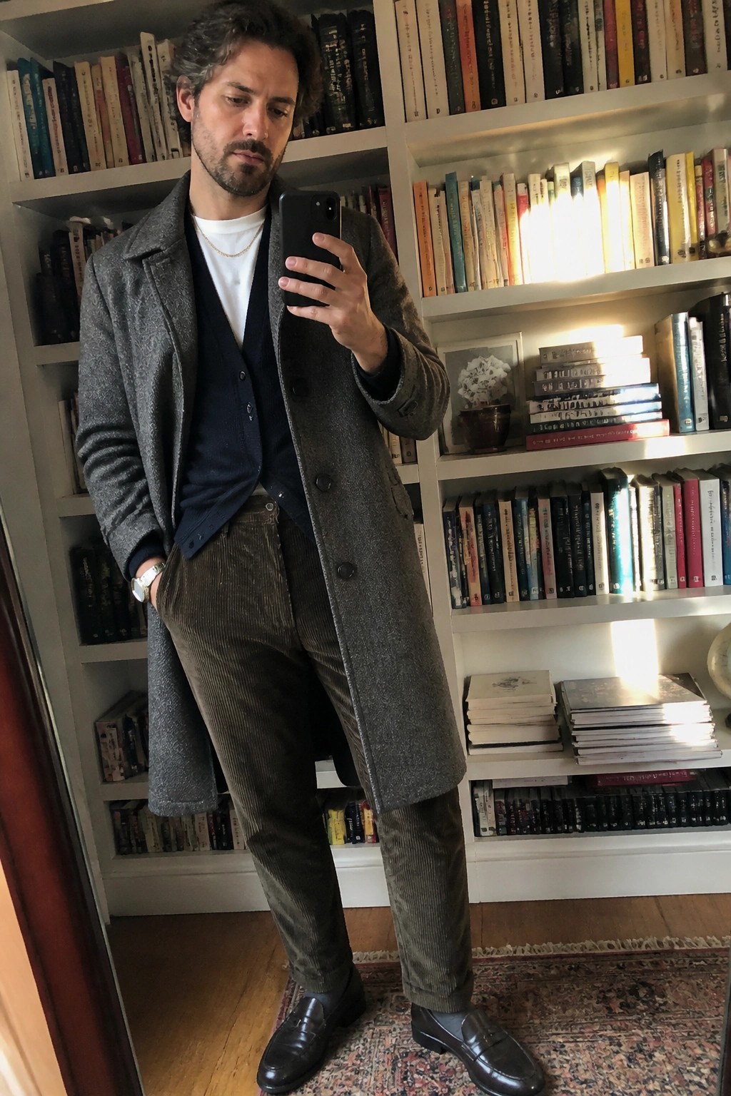 Man in gray overcoat, navy cardigan, and brown corduroy pants taking mirror selfie