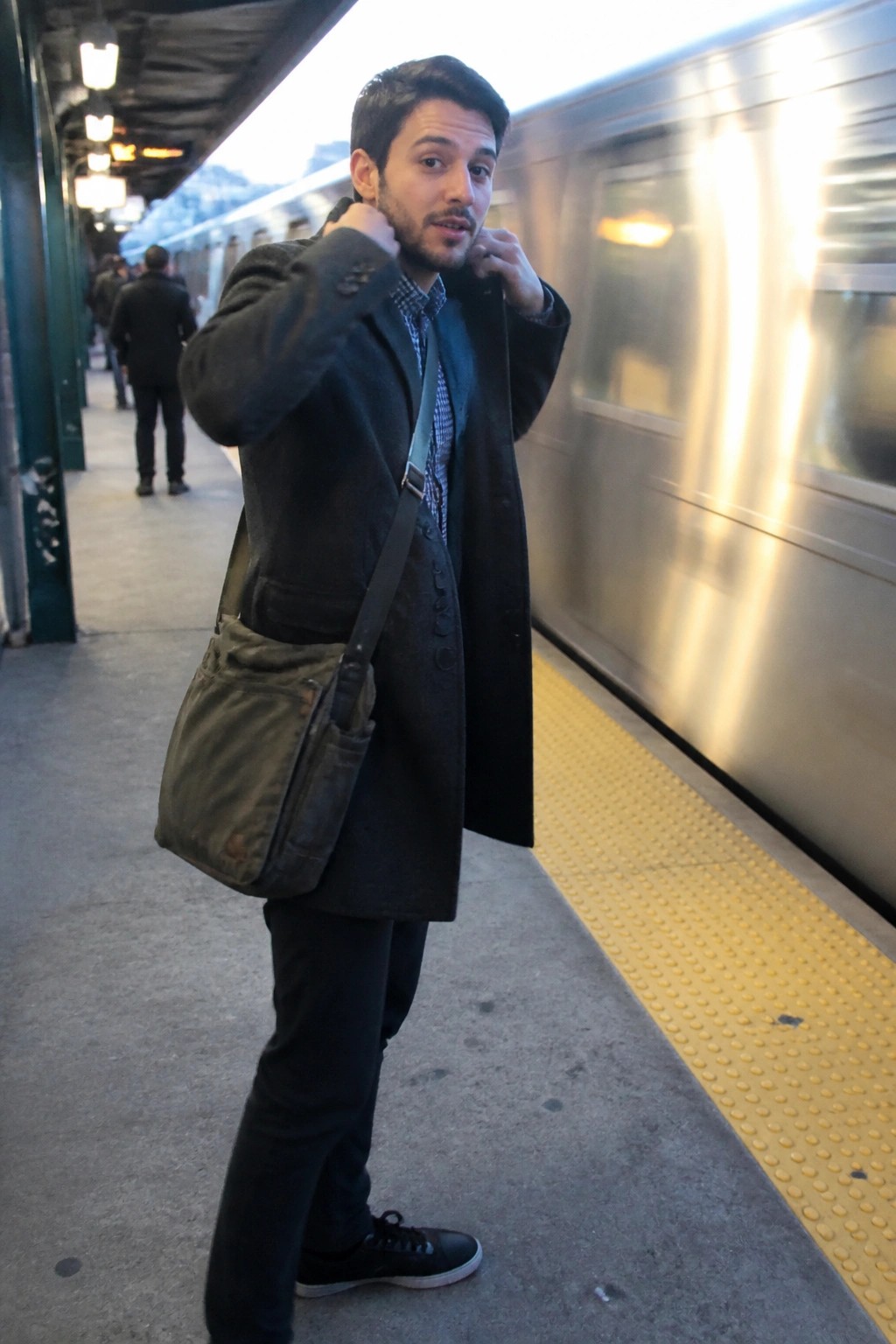 Man in dark coat with green bag on subway platform beside blurred train