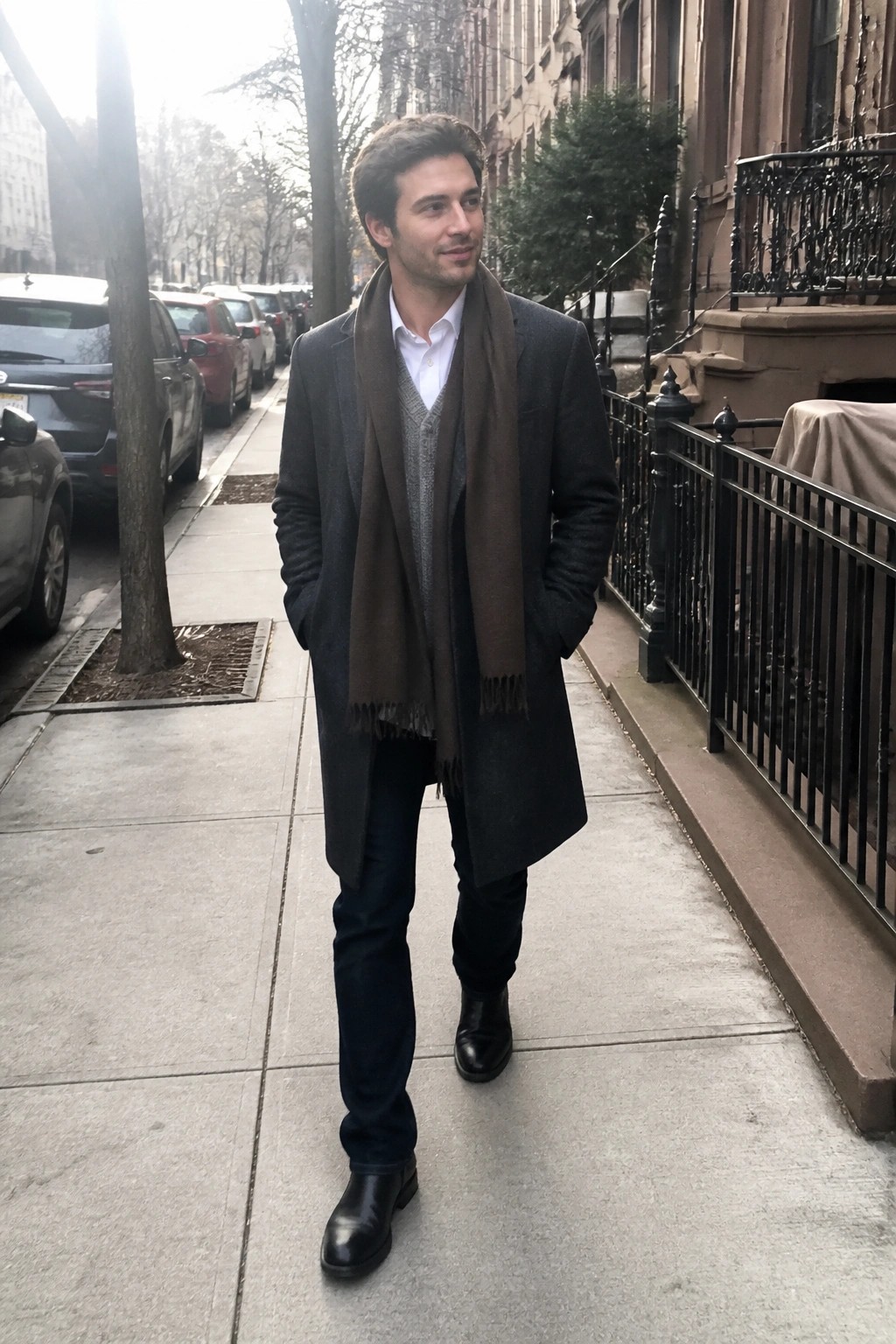 Man in dark overcoat and brown scarf stands on city sidewalk with hands in pockets