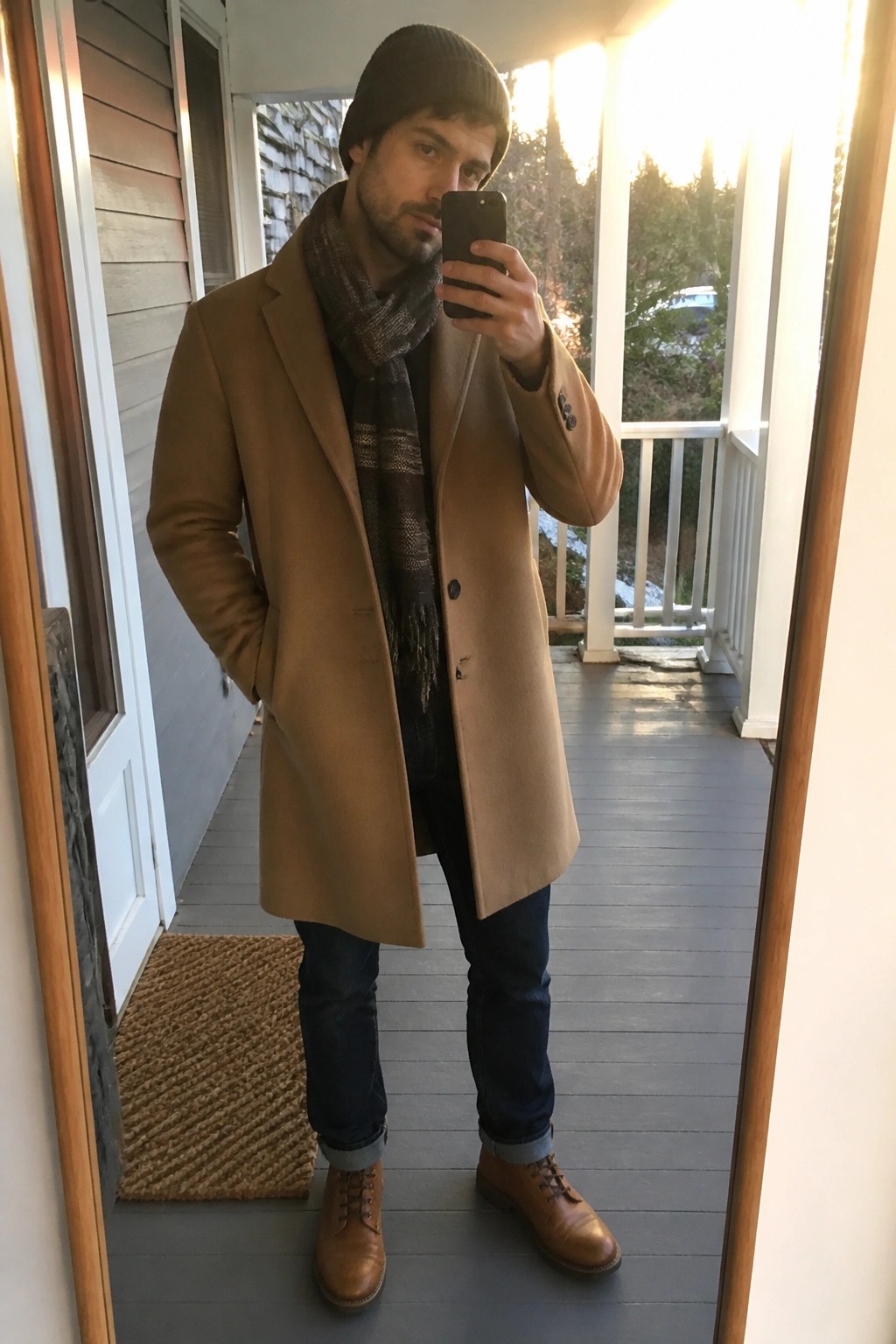 Man in tan coat and beanie taking mirror selfie on wooden porch at sunset
