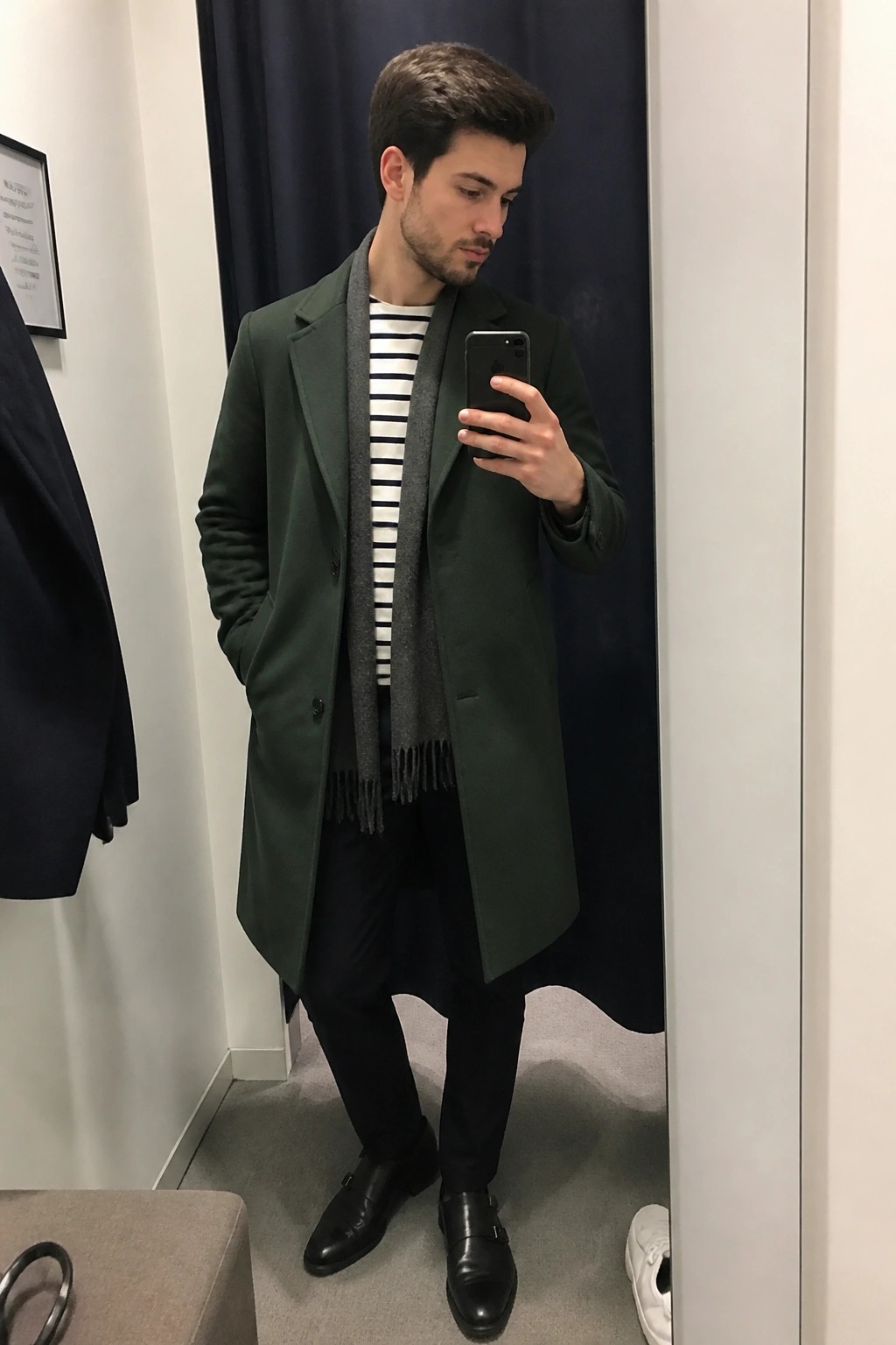 Man in dark green overcoat and gray scarf taking mirror selfie with phone