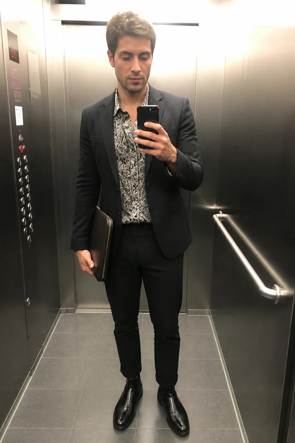 Man in black blazer and patterned shirt taking phone selfie in elevator holding portfolio