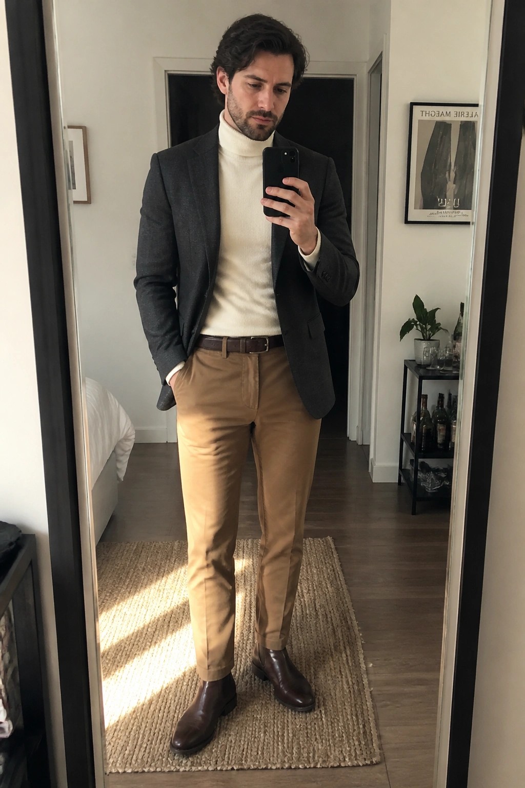 Man in gray blazer and cream turtleneck taking mirror selfie with phone