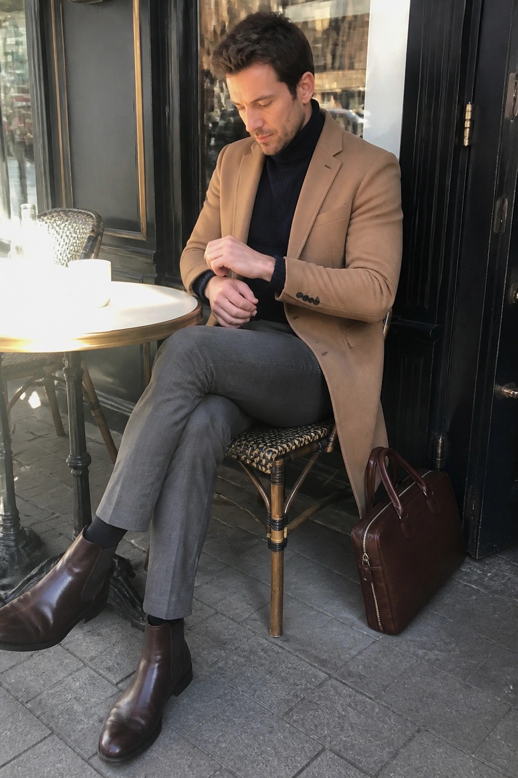 Man in tan overcoat and gray trousers checks watch at cafe with brown briefcase