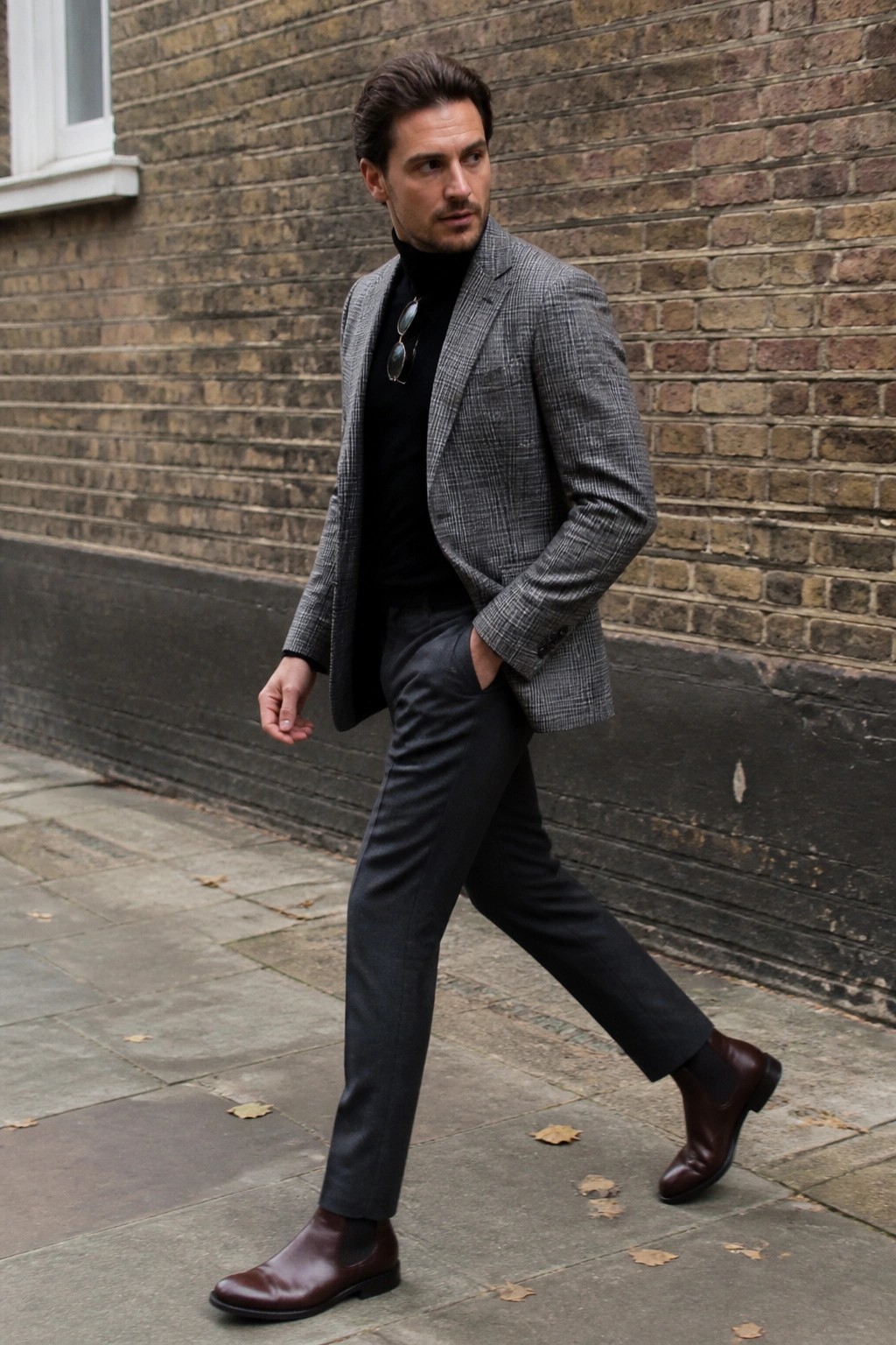 Man in gray plaid blazer over black turtleneck walks on paved sidewalk in brown boots