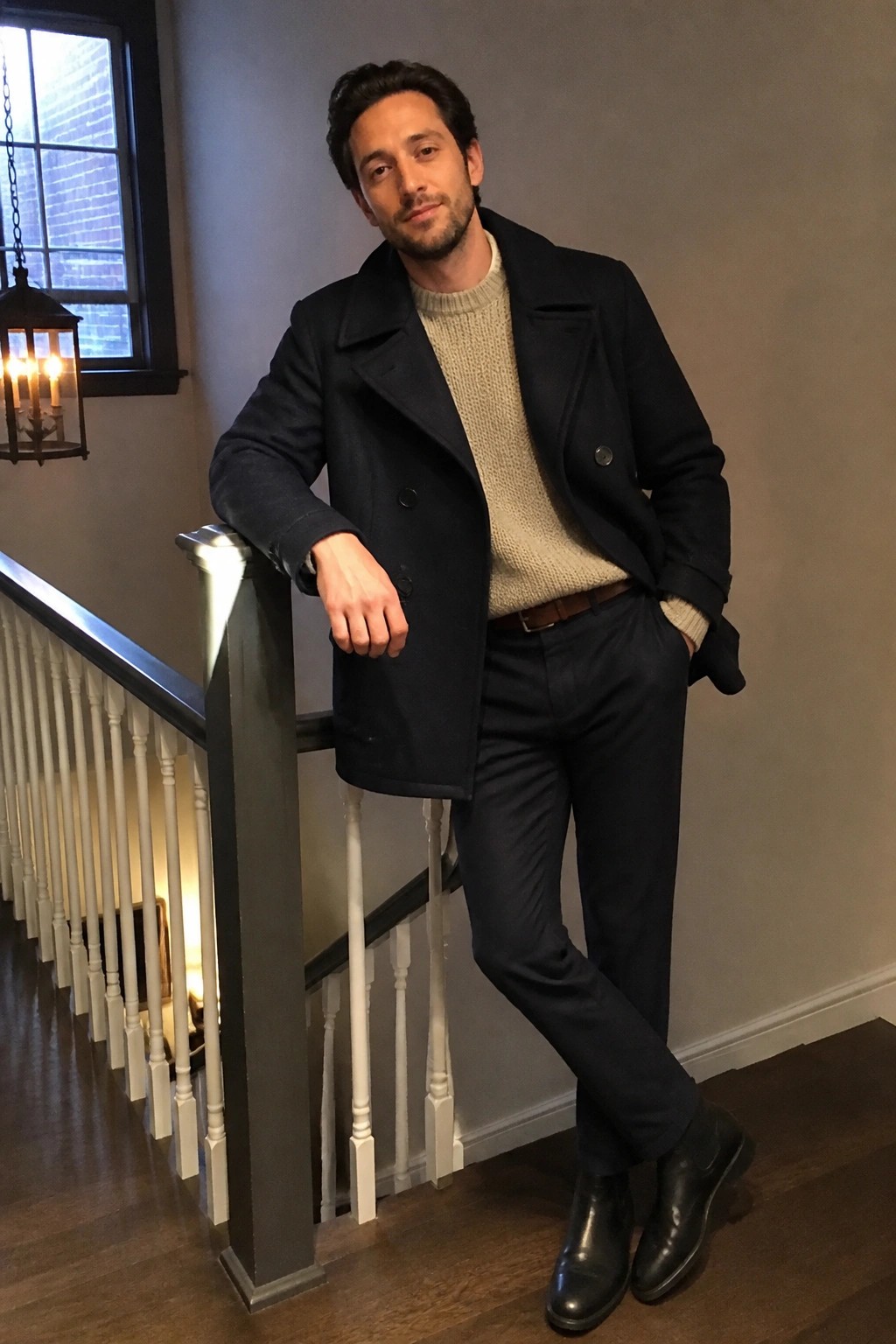 Man in navy peacoat and beige knit sweater leaning on staircase railing
