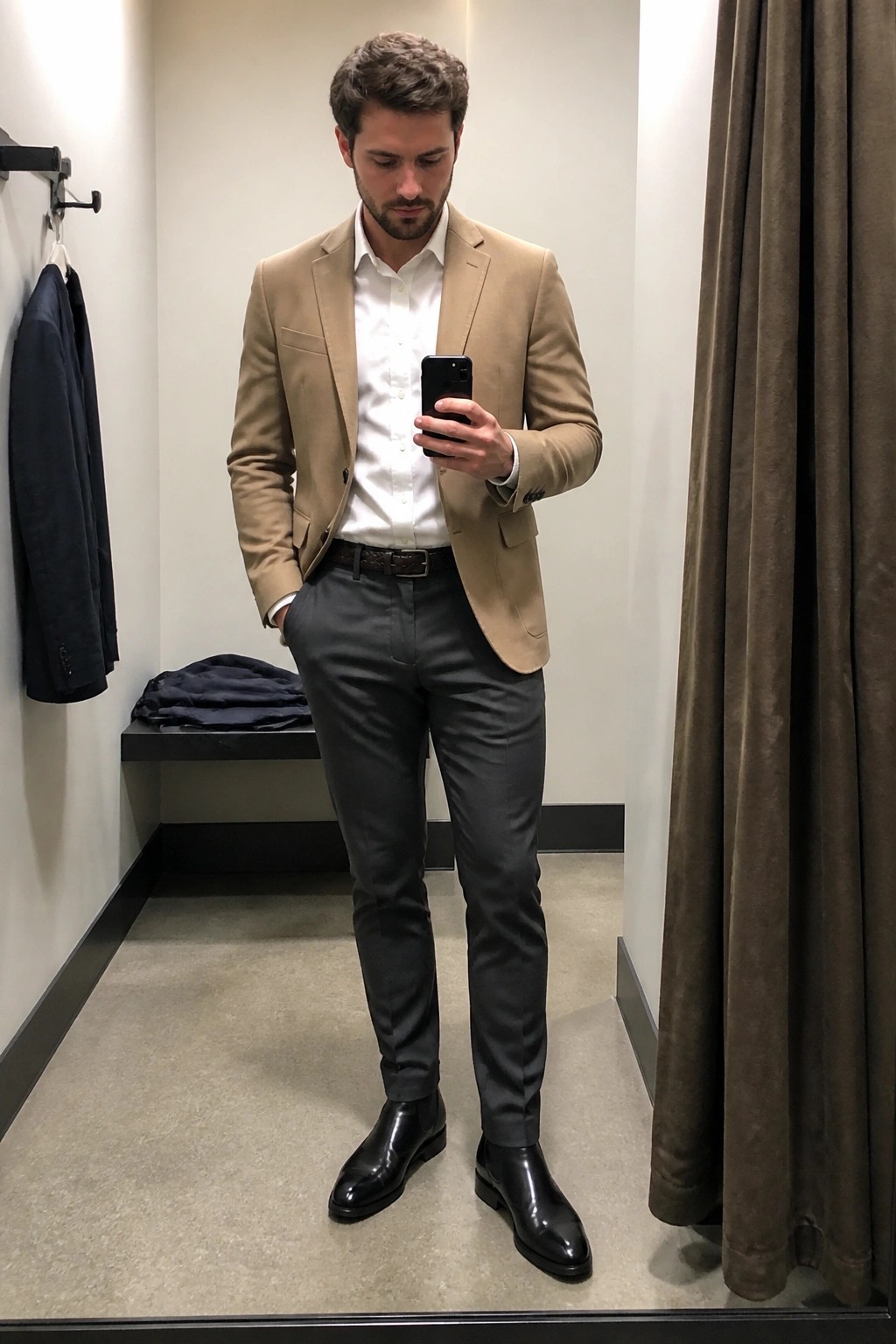 Man in beige blazer and gray pants taking mirror selfie in fitting room