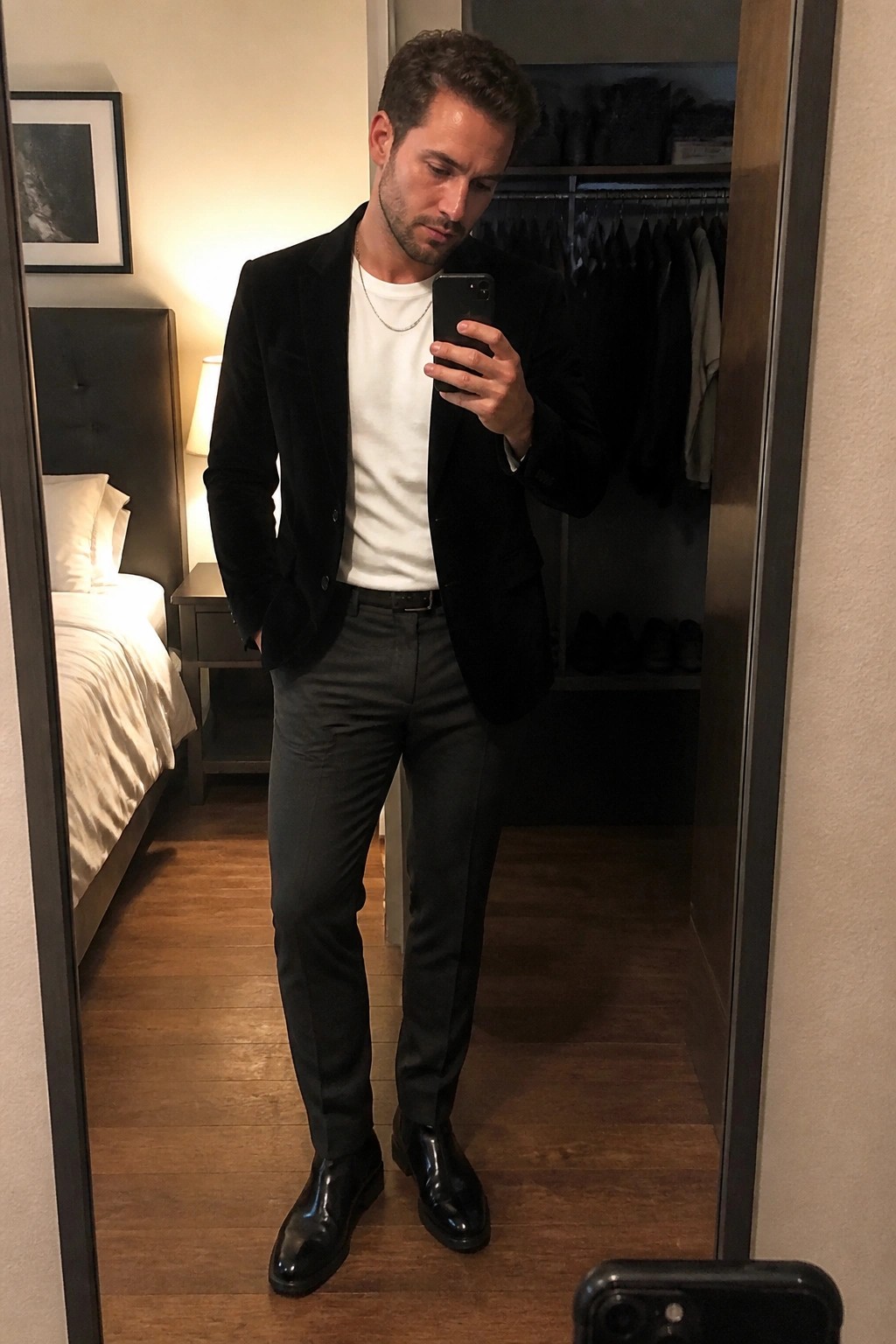 Man in black velvet blazer, white tee, and gray pants taking mirror selfie in bedroom