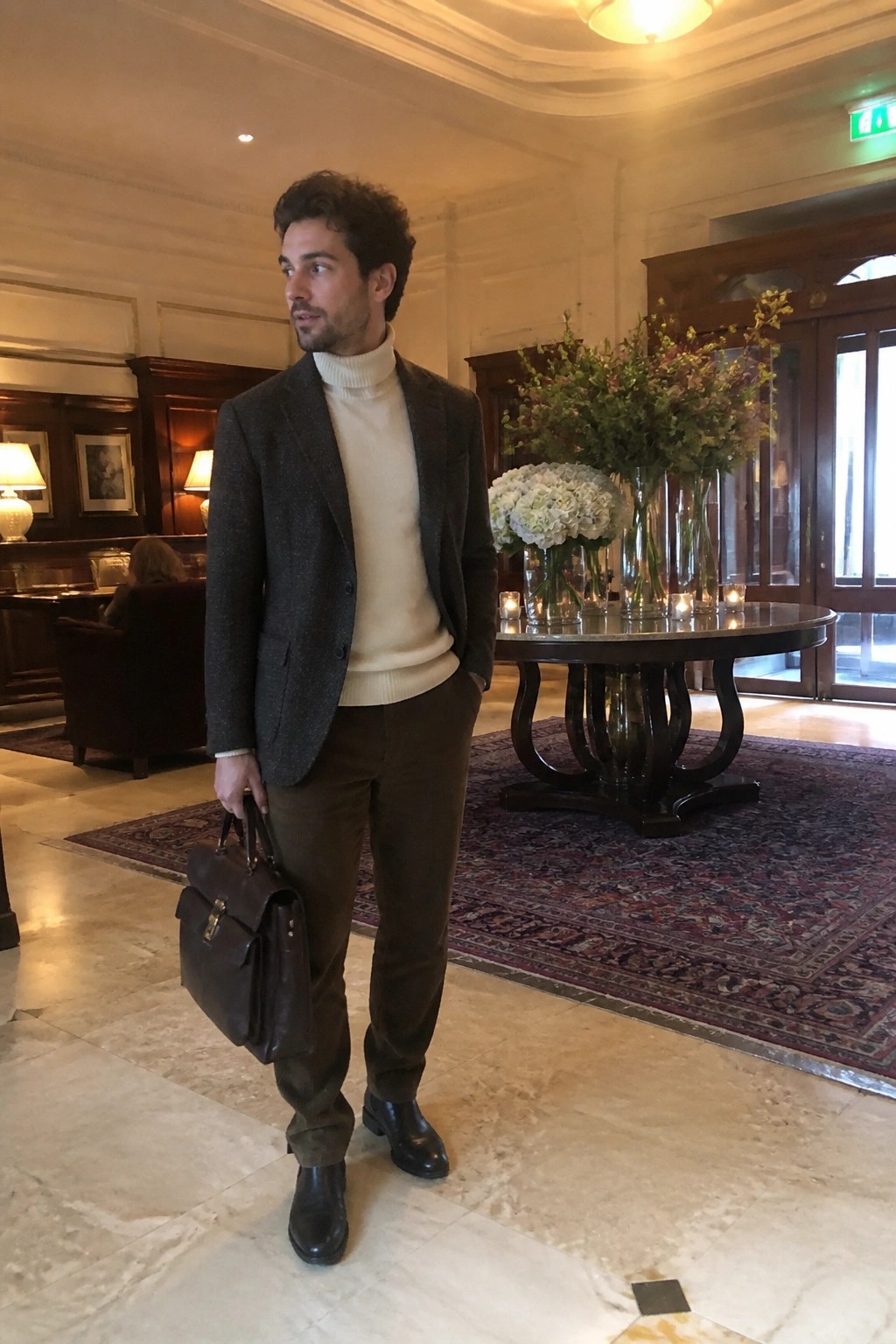 Man in tweed blazer and turtleneck holds briefcase in elegant marble lobby