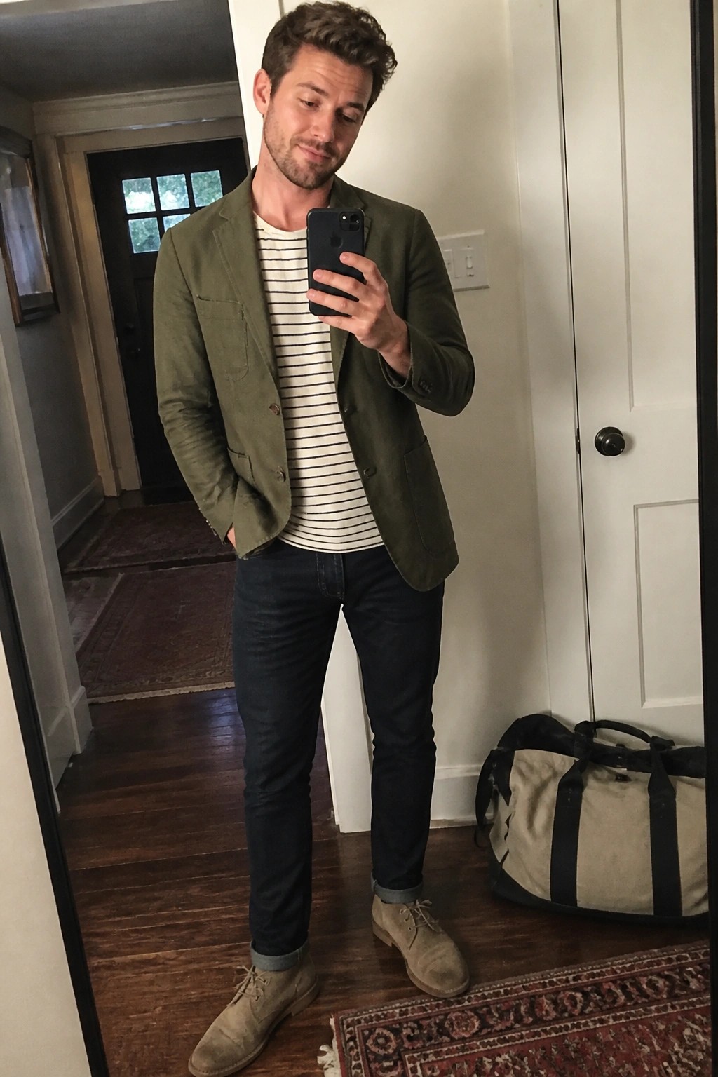 Man wearing an unstructured olive green blazer over a white and navy striped t-shirt, paired with dark blue cuffed jeans and beige suede chukka boots, holding a phone in a mirror selfie.