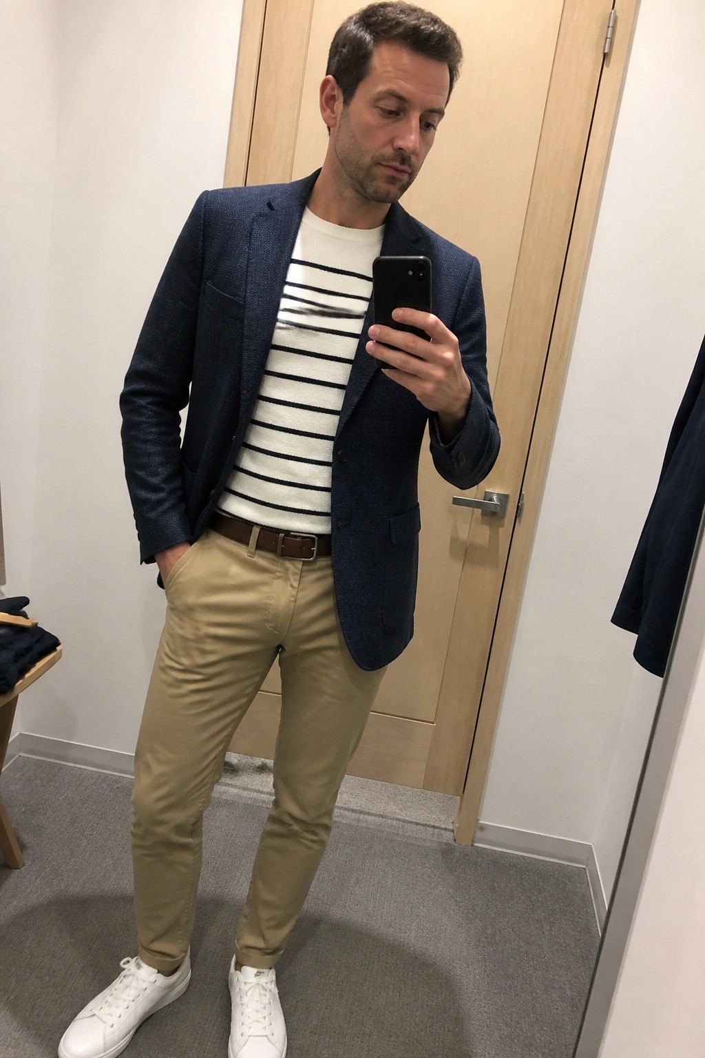 Man in navy blazer over striped sweater with khaki pants and white sneakers holds phone.