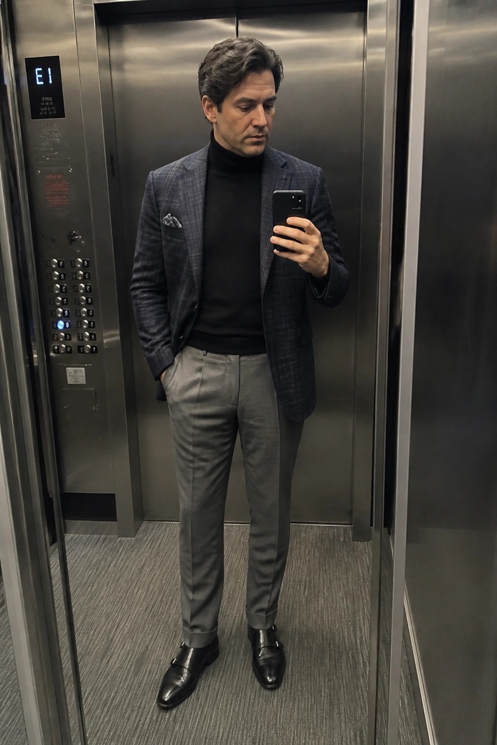 Man in plaid blazer and turtleneck holding phone inside elevator