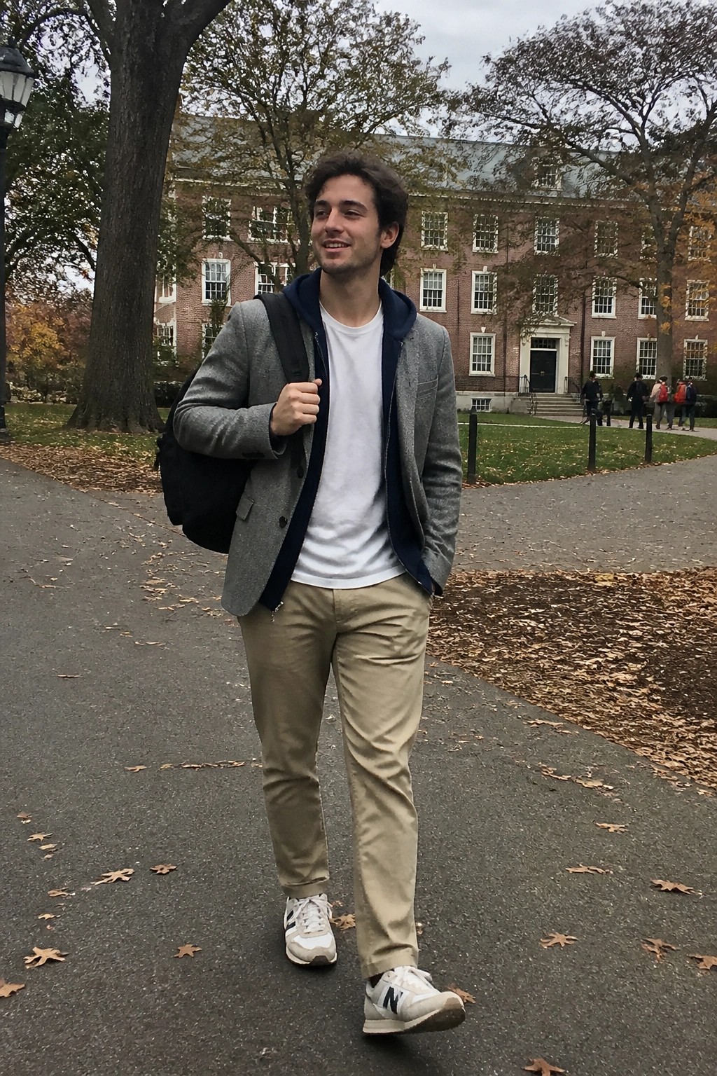 Smiling young man in gray blazer over hoodie walks on leafy campus path with backpack