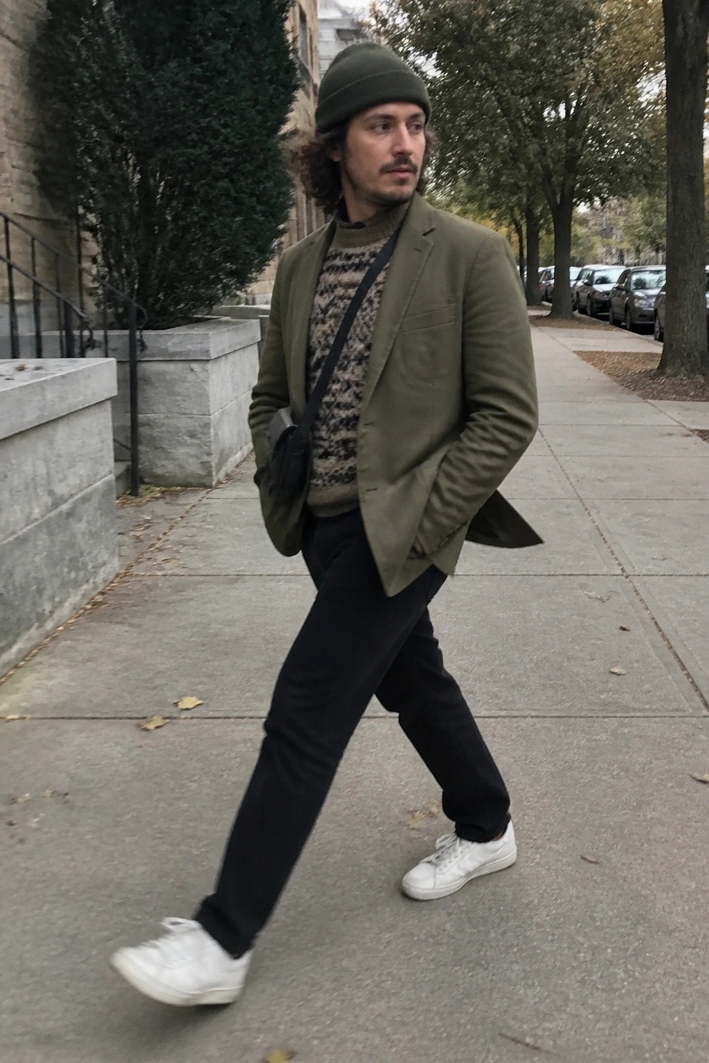 Man in olive blazer and beanie walks city sidewalk with crossbody bag