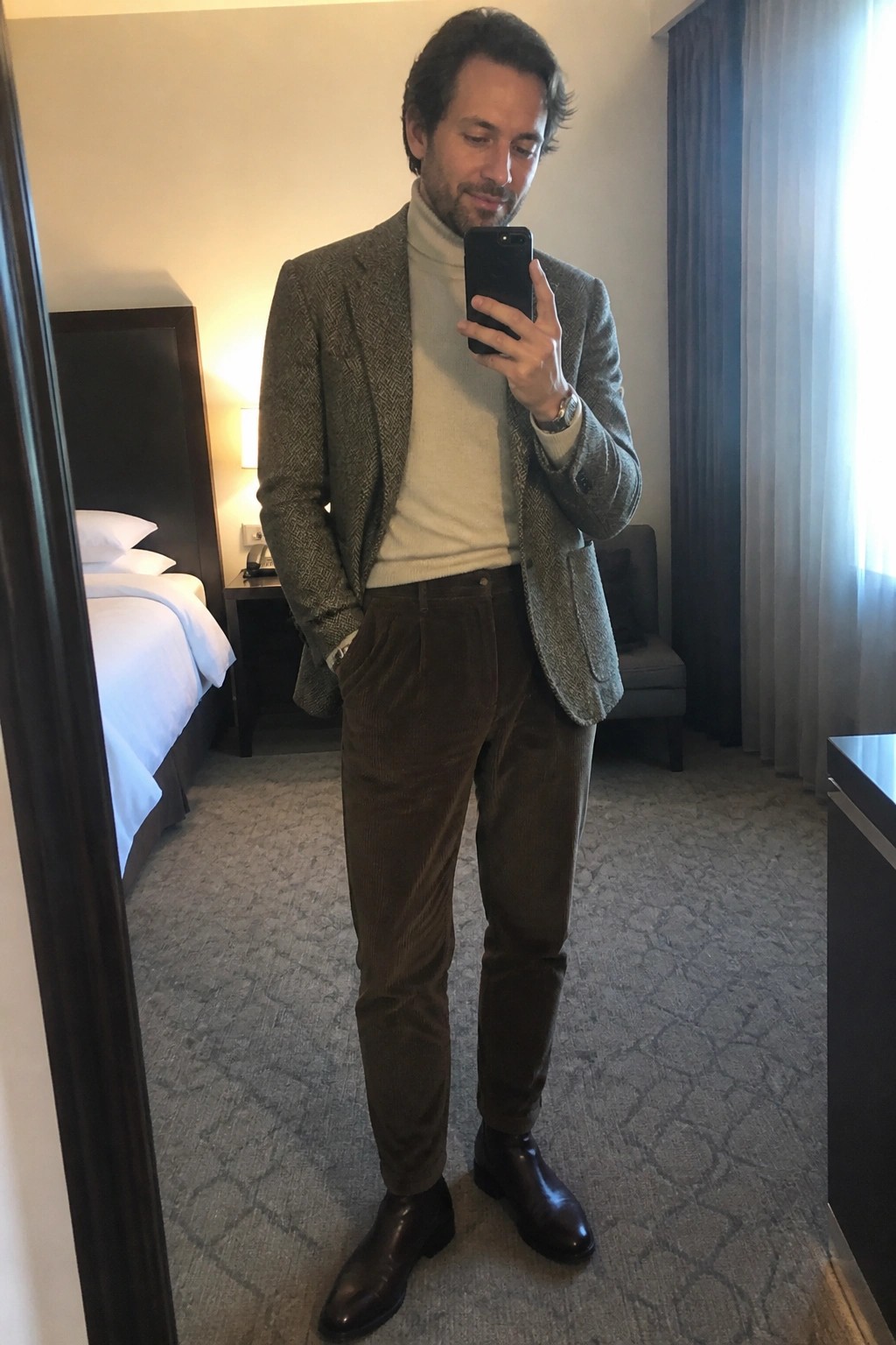 Man in tweed blazer and corduroy pants taking mirror selfie in hotel room