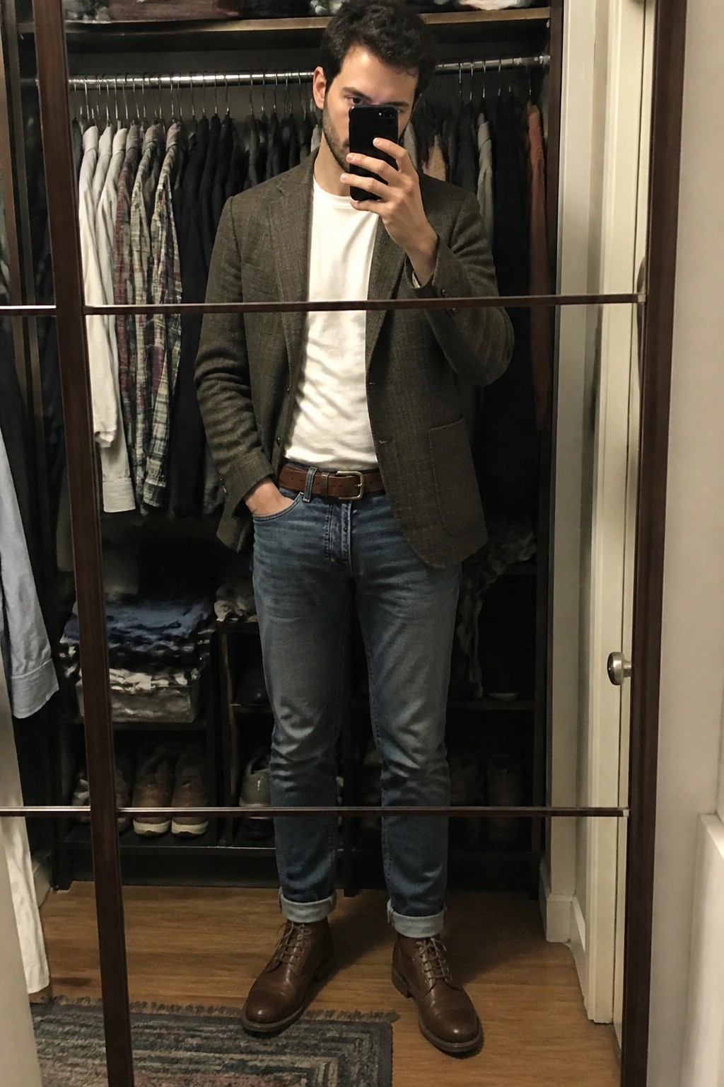 Man in brown blazer, white tee, and jeans taking mirror selfie in closet