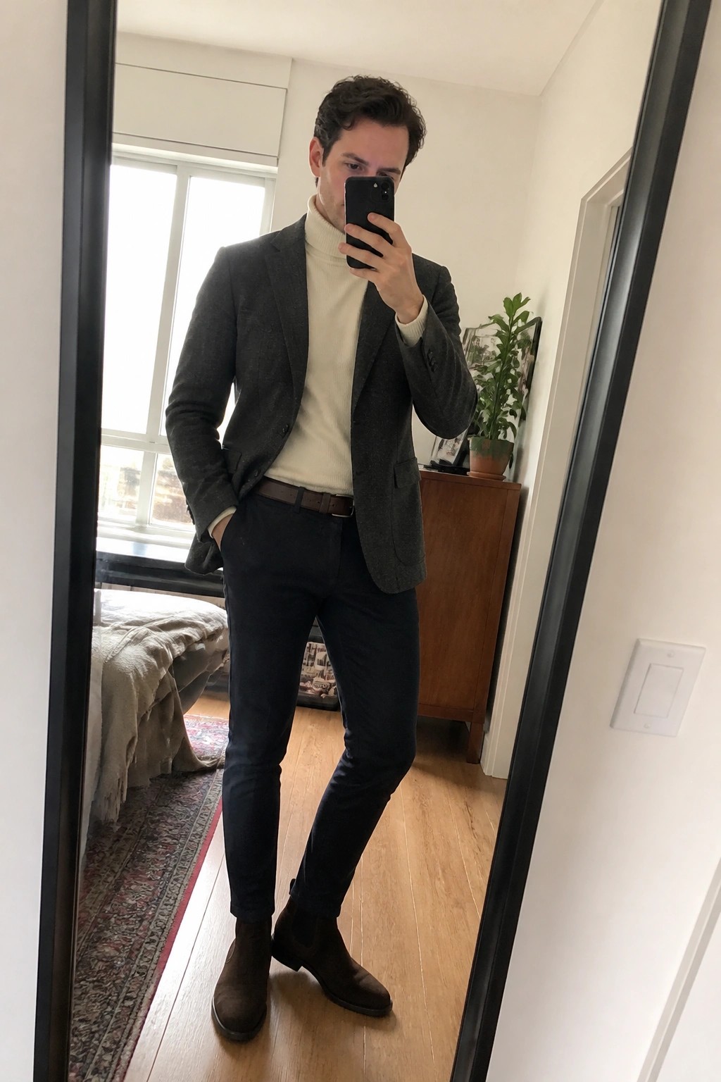 Man in gray blazer and cream turtleneck taking mirror selfie in bedroom