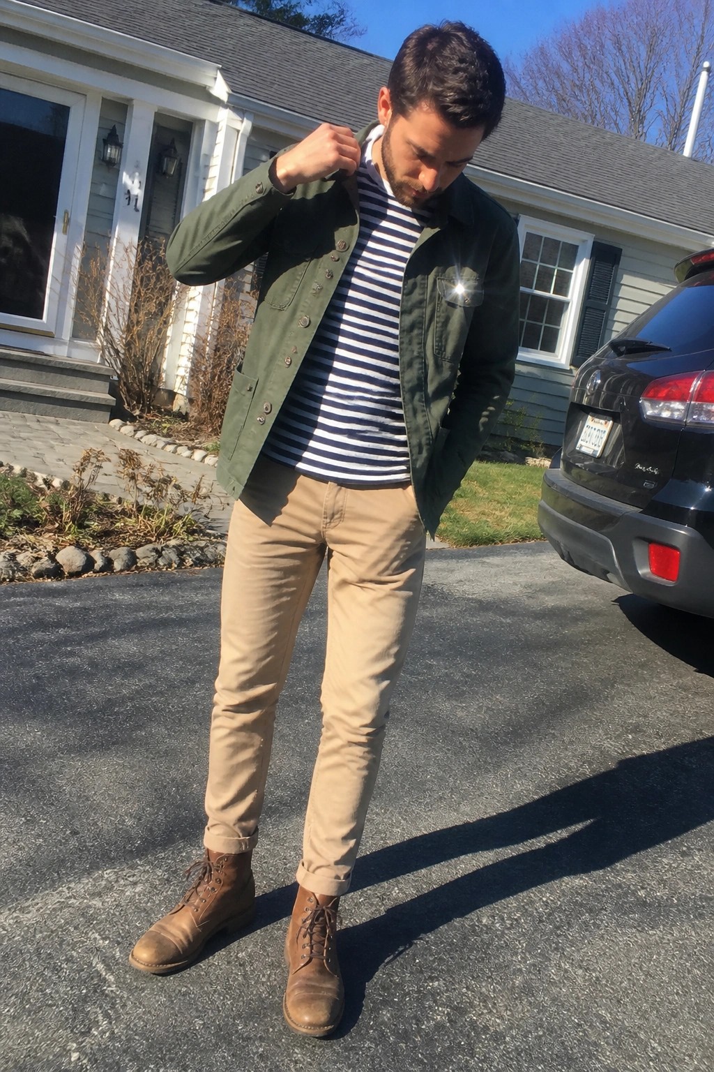 Man in green jacket over striped shirt adjusts collar on sunny driveway with black SUV nearby.