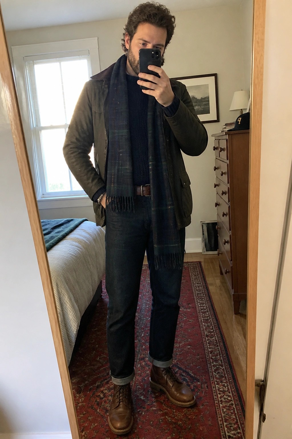 Man in green jacket and plaid scarf taking mirror selfie in bedroom