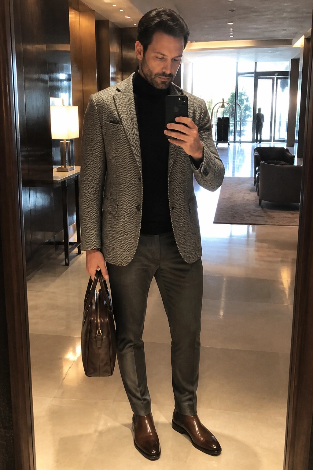 Man wearing grey herringbone blazer over black turtleneck with grey trousers and brown leather boots while holding a matching leather bag.