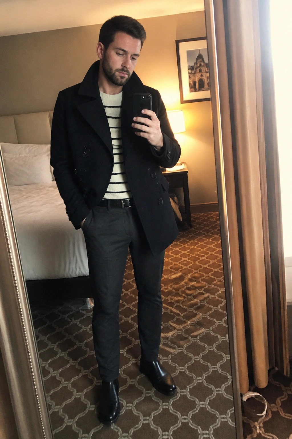 Man in black peacoat and striped sweater taking mirror selfie in hotel room