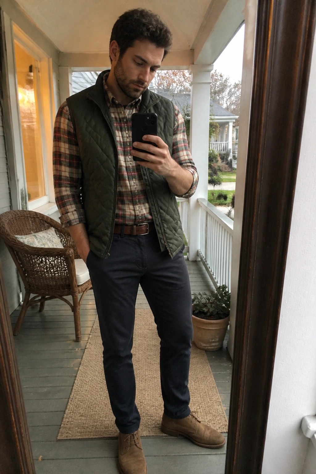 Man in green quilted vest over plaid shirt takes phone selfie on porch.