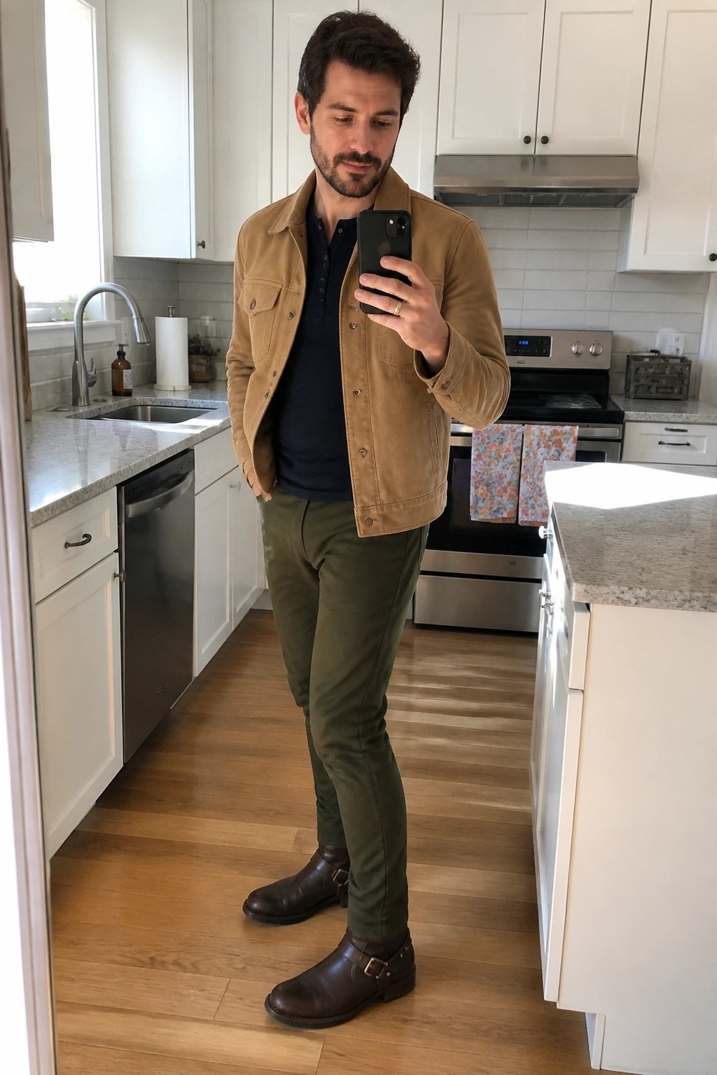 Man in tan suede jacket and olive pants taking phone selfie in kitchen