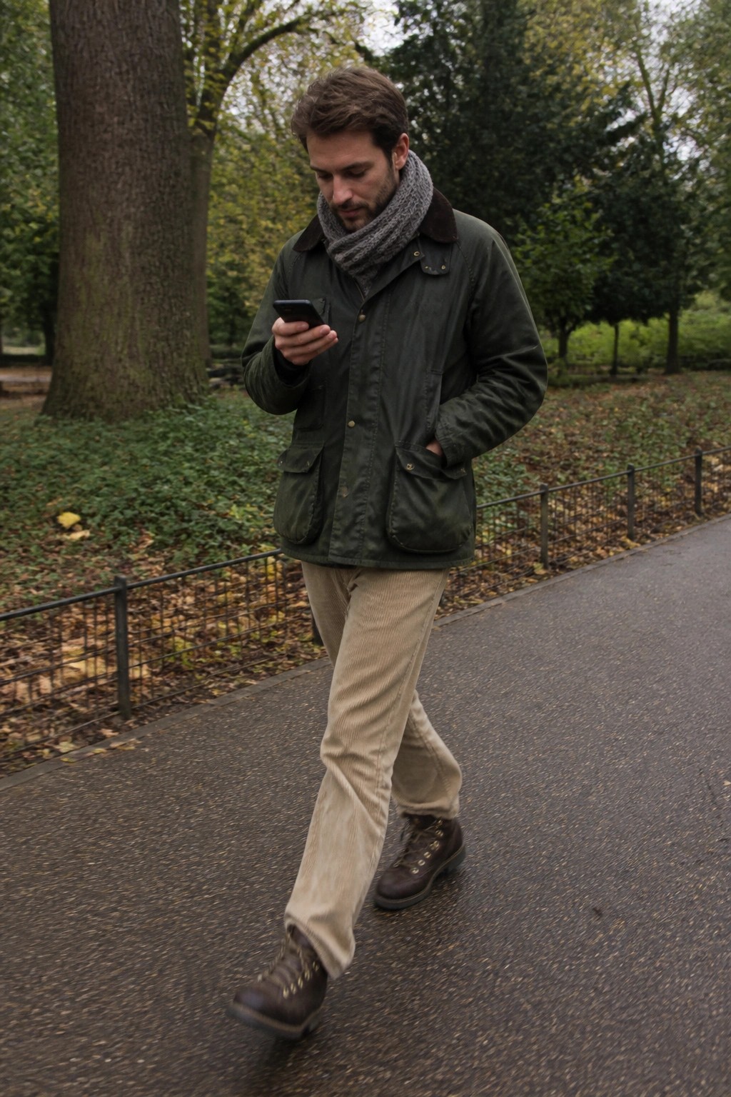 Man in green waxed field jacket, gray knit scarf, beige corduroy trousers, and brown leather boots walking on a path while checking his phone.