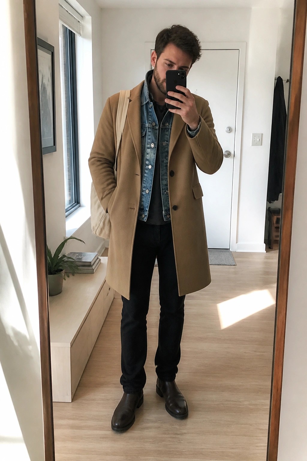 Man in tan overcoat over denim jacket taking mirror selfie in apartment hallway
