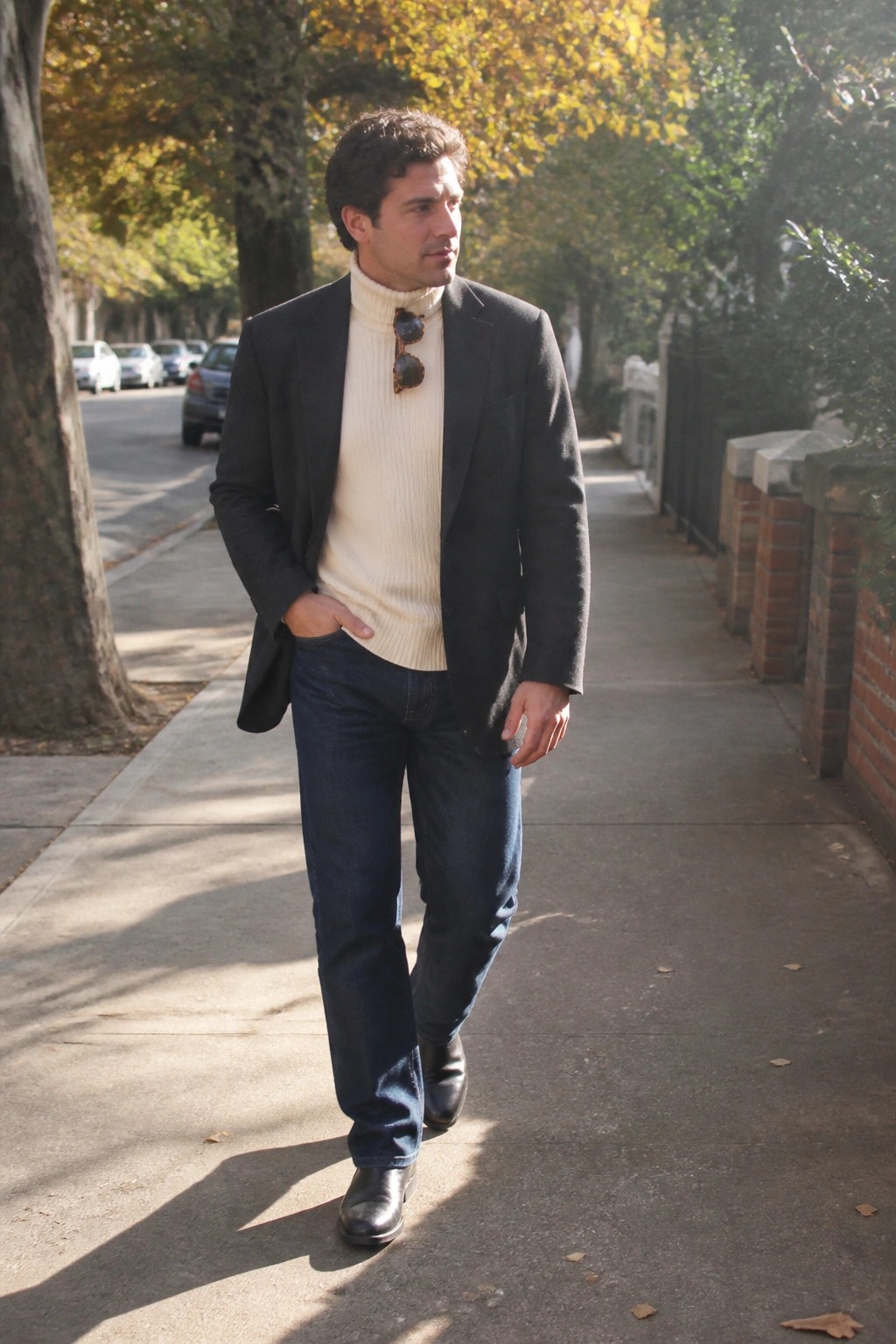 Man in dark blazer over cream turtleneck with sunglasses walks on autumn sidewalk
