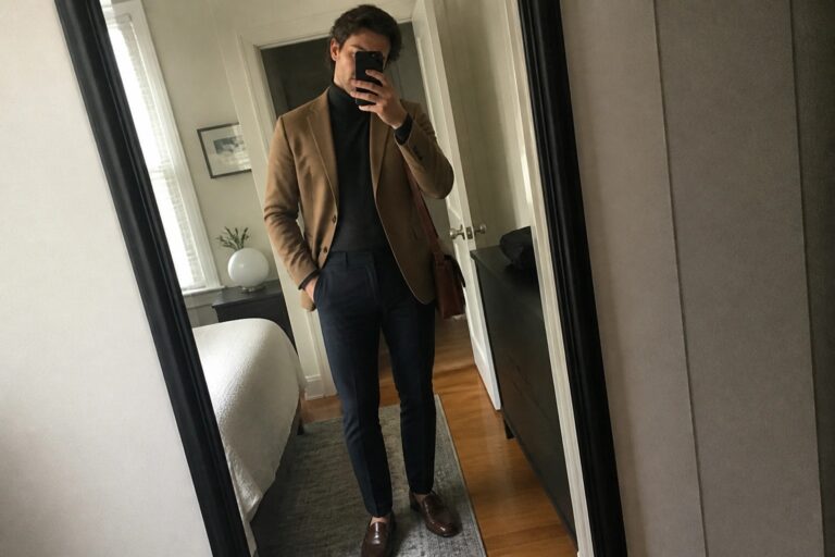 Man in tan blazer over black turtleneck with navy pants taking bedroom mirror selfie