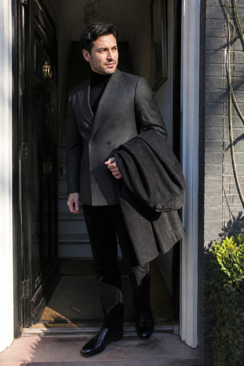Man in gray double-breasted suit and black turtleneck holding overcoat in doorway