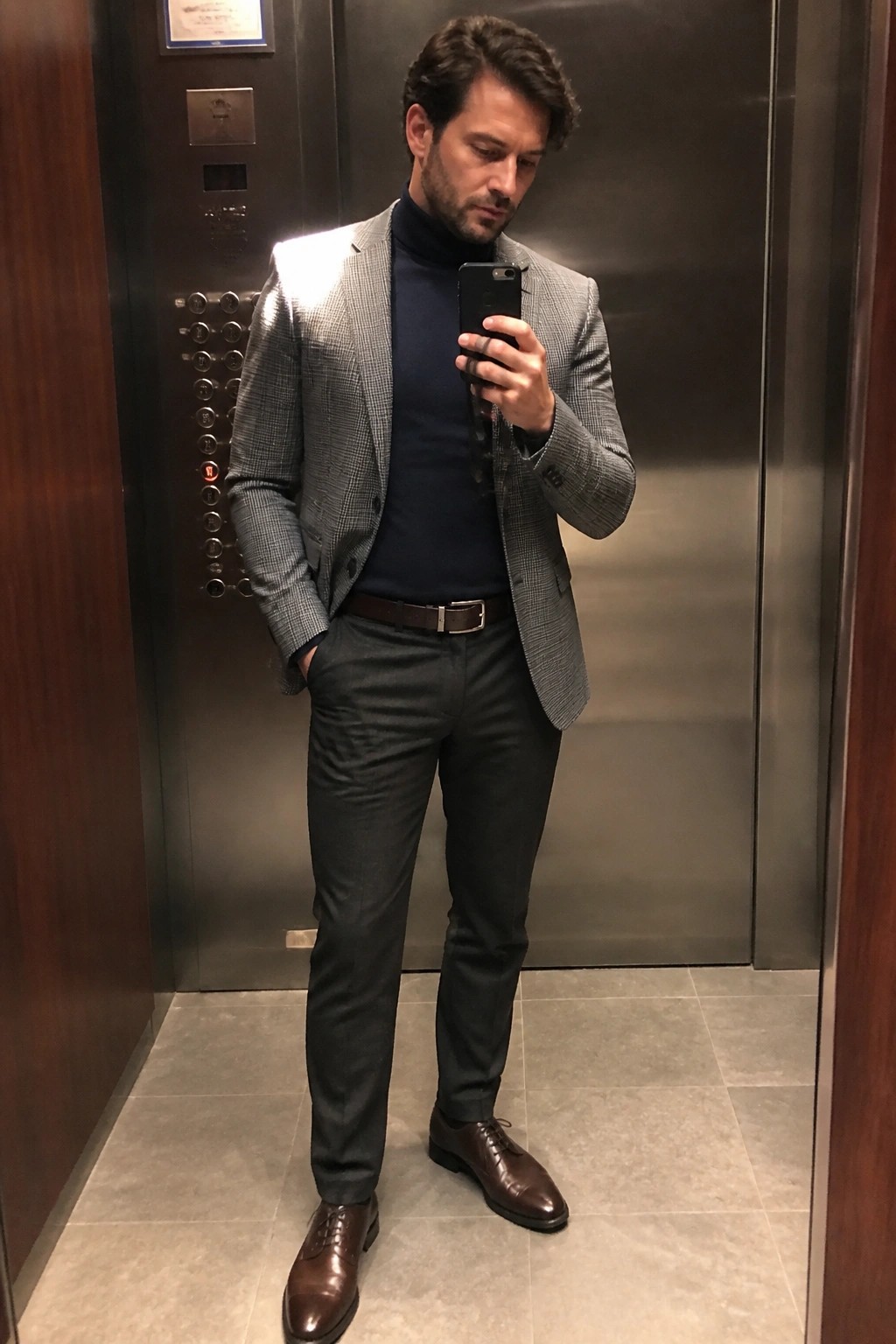 Man in checkered blazer and navy turtleneck taking selfie in elevator