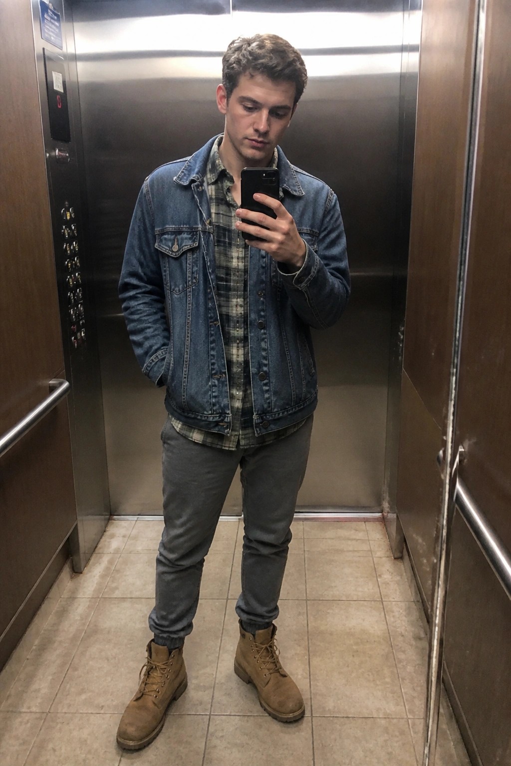 Young man in denim jacket over plaid shirt takes selfie in elevator