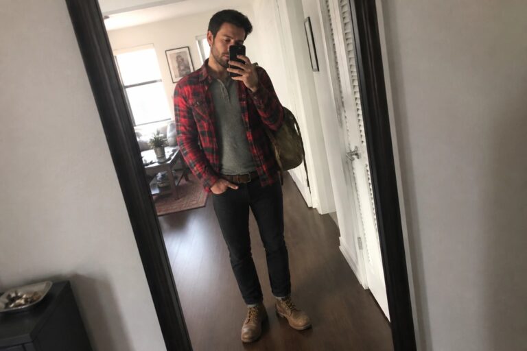 Man in red plaid shirt over gray top with jeans and boots taking mirror selfie