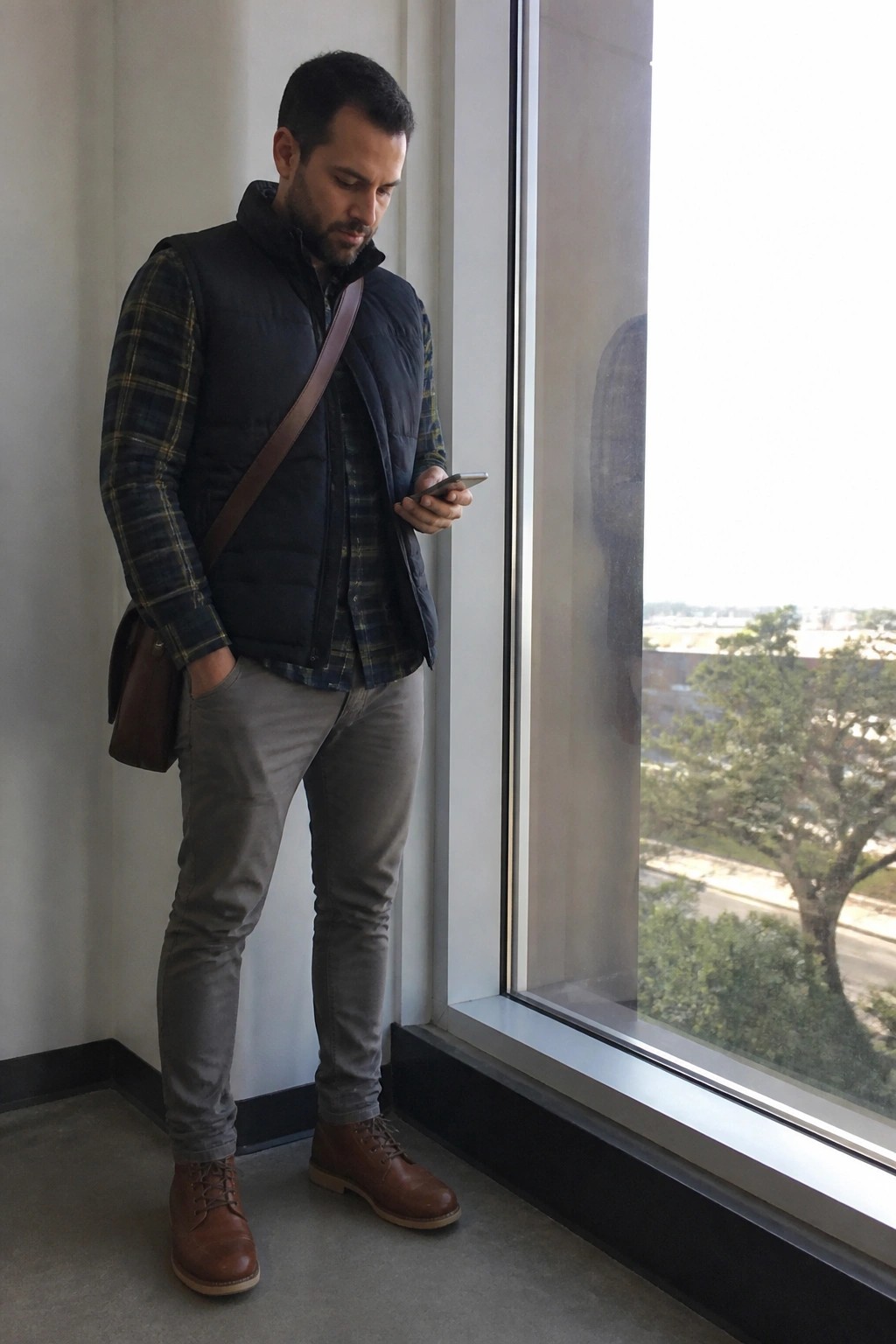Man standing by window wearing plaid flannel shirt under navy puffer vest, gray pants, brown leather work boots, and brown leather crossbody bag while looking at phone.