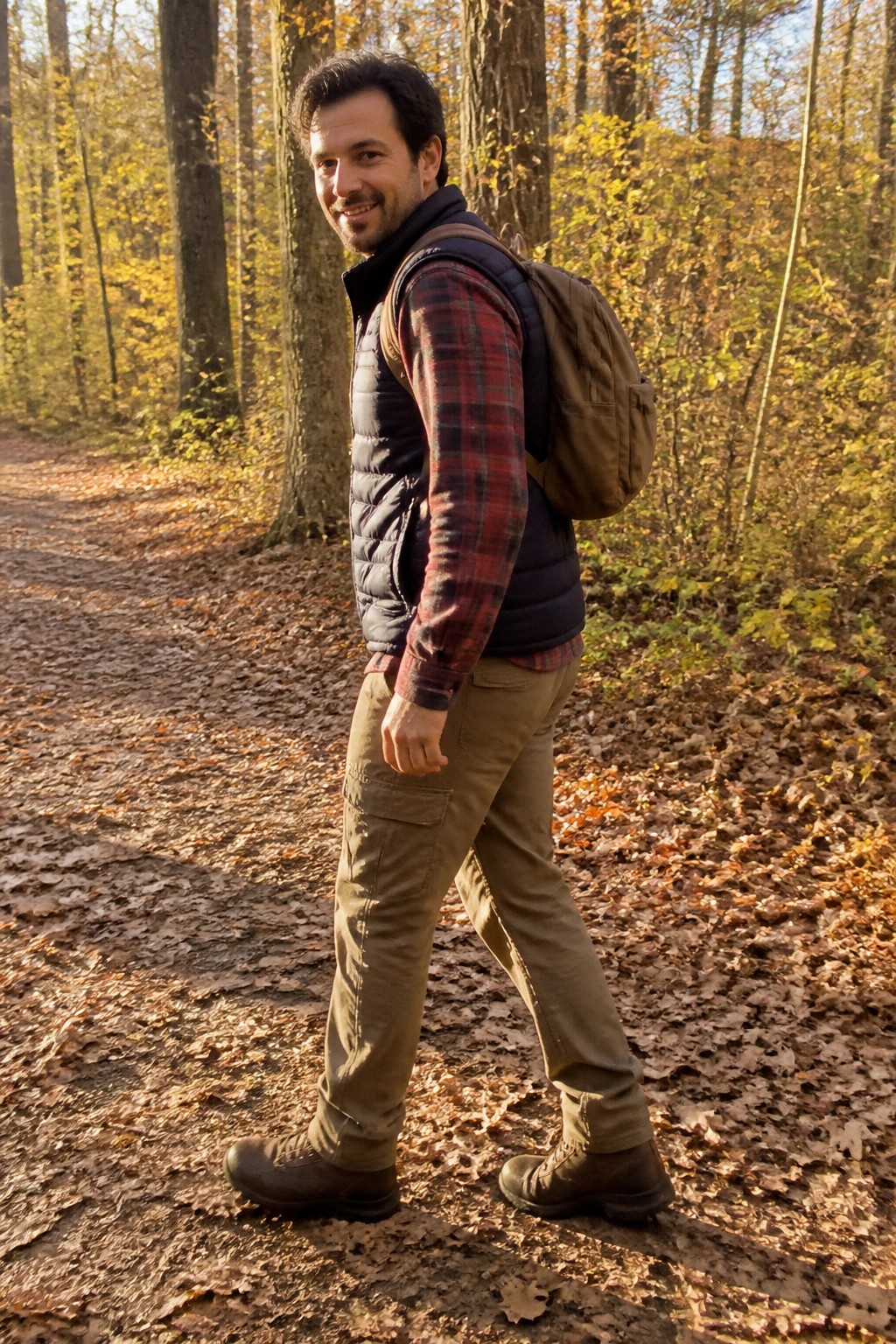Man in red plaid shirt and vest with backpack on autumn forest path