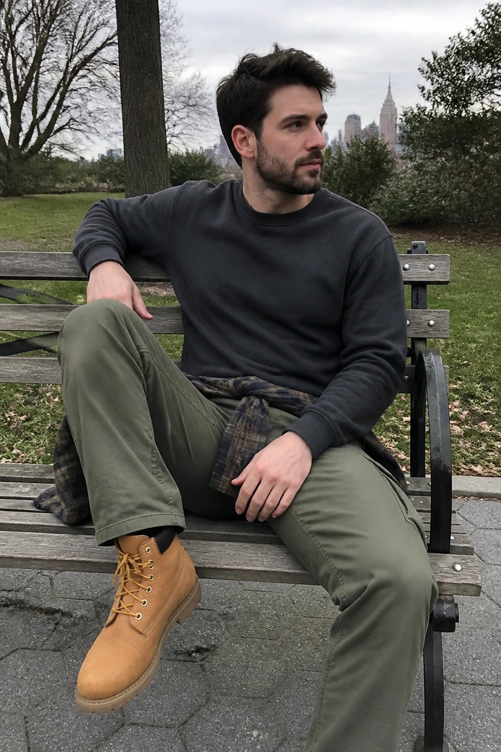 Man in gray sweatshirt and olive pants sits on park bench with city skyline behind