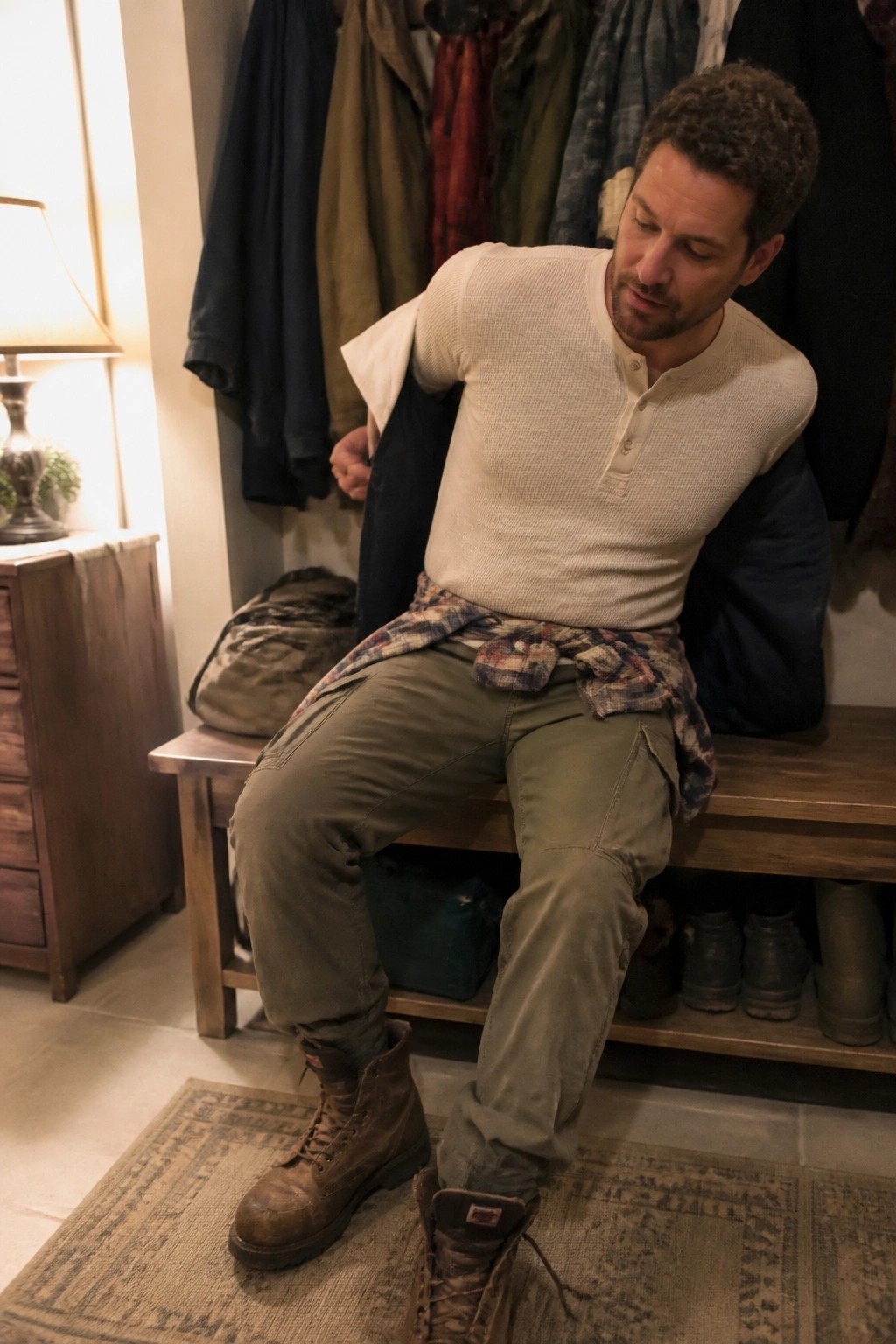 Man in beige henley and cargo pants removing brown boot on wooden bench