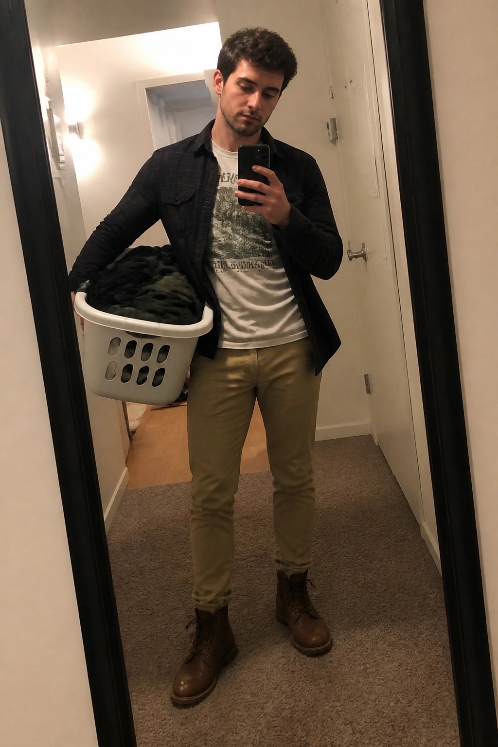 Man in plaid shirt holds laundry basket during mirror selfie in hallway