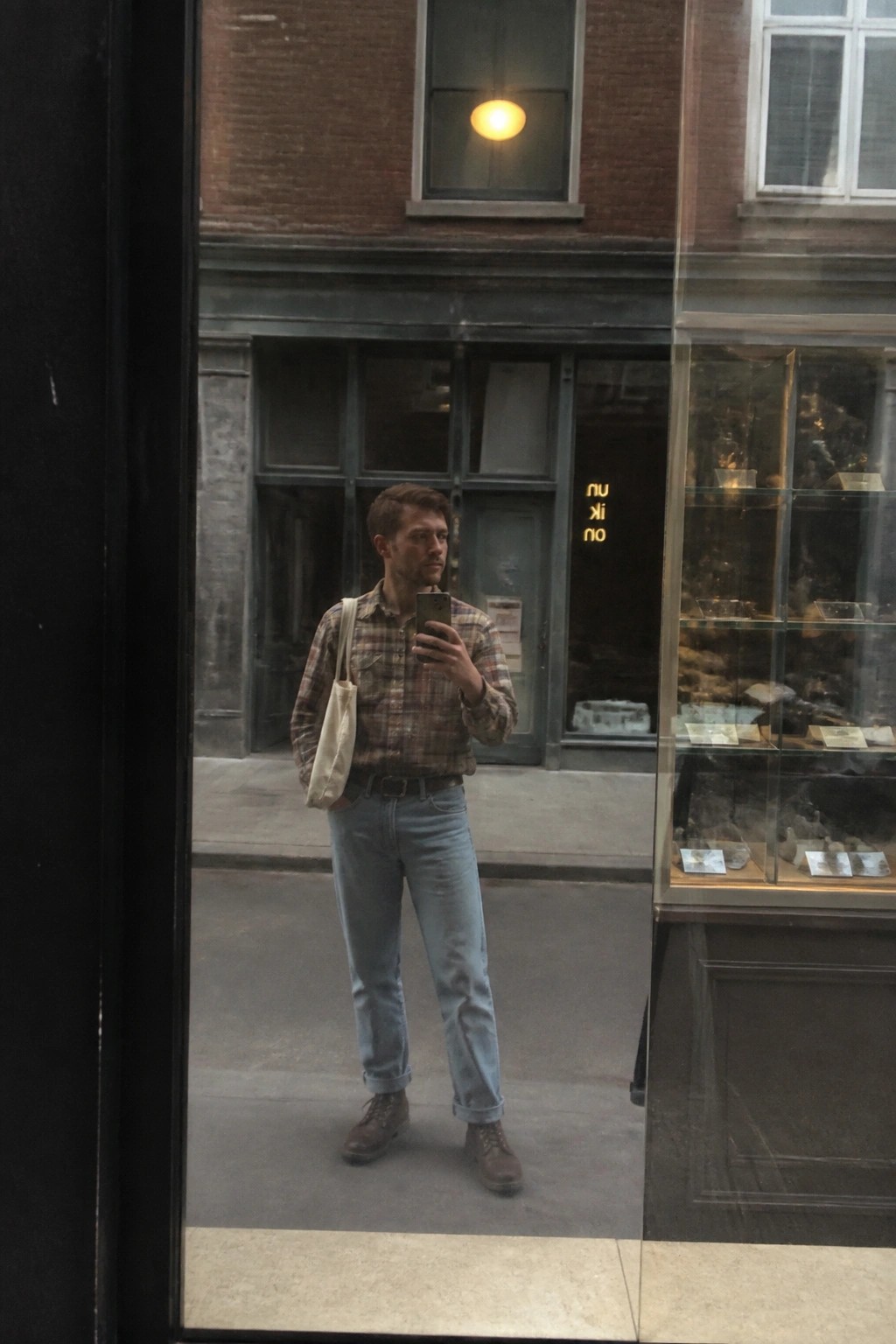 Man wearing brown and green plaid flannel shirt tucked into light blue cuffed jeans with brown work boots and beige canvas tote bag standing in front of a storefront window while holding a phone.
