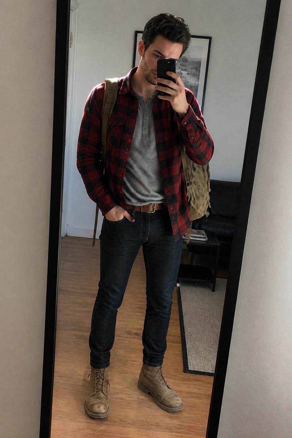 Man in red plaid shirt over gray top with jeans and boots taking mirror selfie