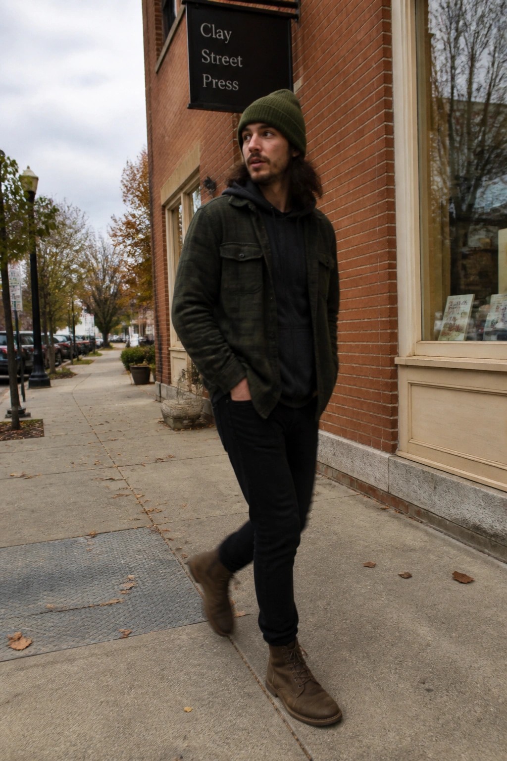 Long-haired man in green beanie and plaid shirt stands on sidewalk by brick building.