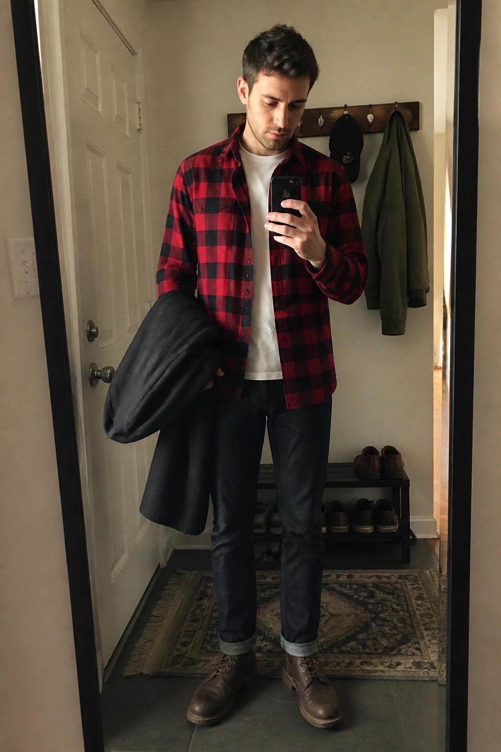 Man in red plaid shirt holding coat takes mirror selfie in entryway