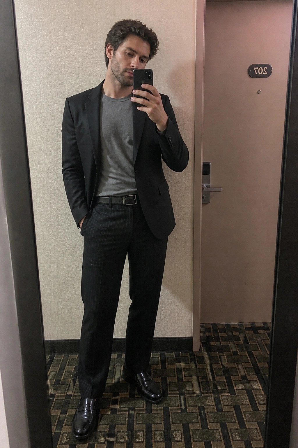 Man in black blazer over gray t-shirt with black pinstripe trousers, black belt, and black loafers standing in front of a door while holding a phone for a mirror selfie.